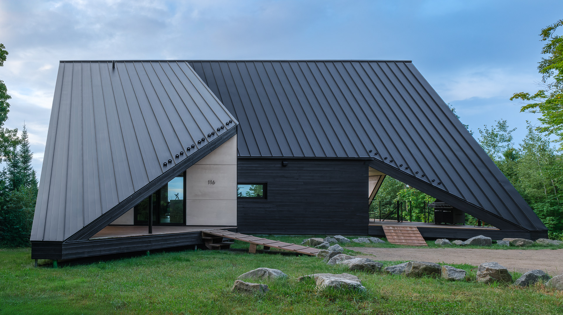Façade d'accès de la Cabine-A à Charlevoix par Bourgeois / Lechasseur de jour. L'imposante toiture métallique asymétrique descend jusqu'au sol rocheux, complétée par un revêtement en bois noir et des rampes d'accès en bois naturel.