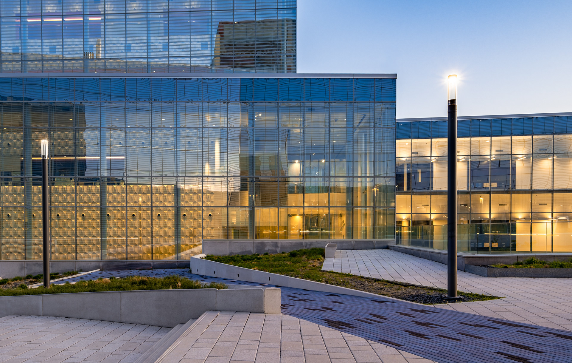 Aménagement extérieur et façade du Complexe des sciences au Campus MIL. Le calepinage contrasté au sol et les emmarchements paysagers bordent le pavillon aux vitrages frittés laissant transparaître les espaces intérieurs éclairés,