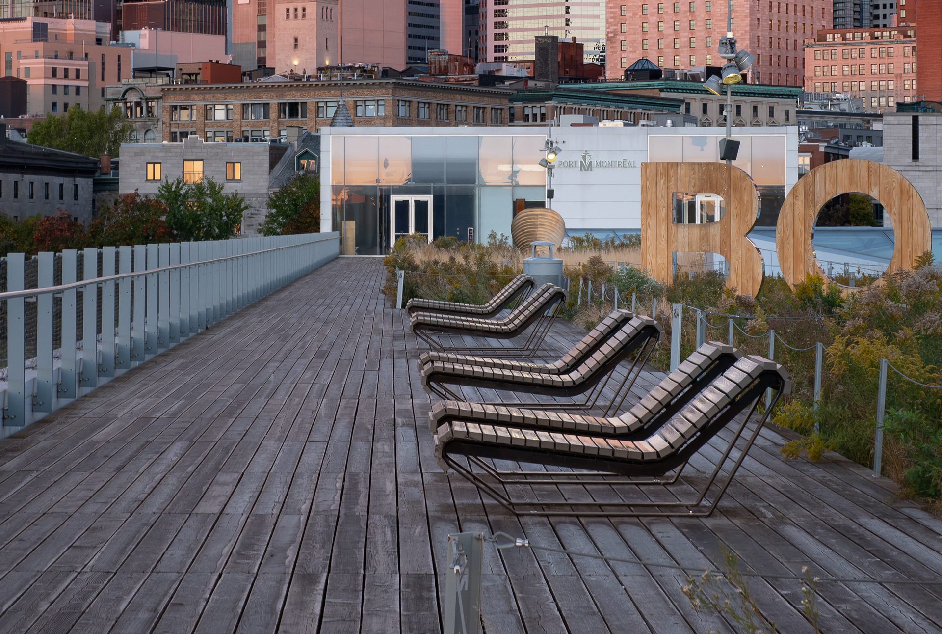 Zone de détente sur la toiture du Grand Quai de Montréal, conçue par NIPPAYSAGE et Provencher_Roy. Des chaises longues ergonomiques font face à la végétation naturalisée, devant le paysage urbain à l'aube.