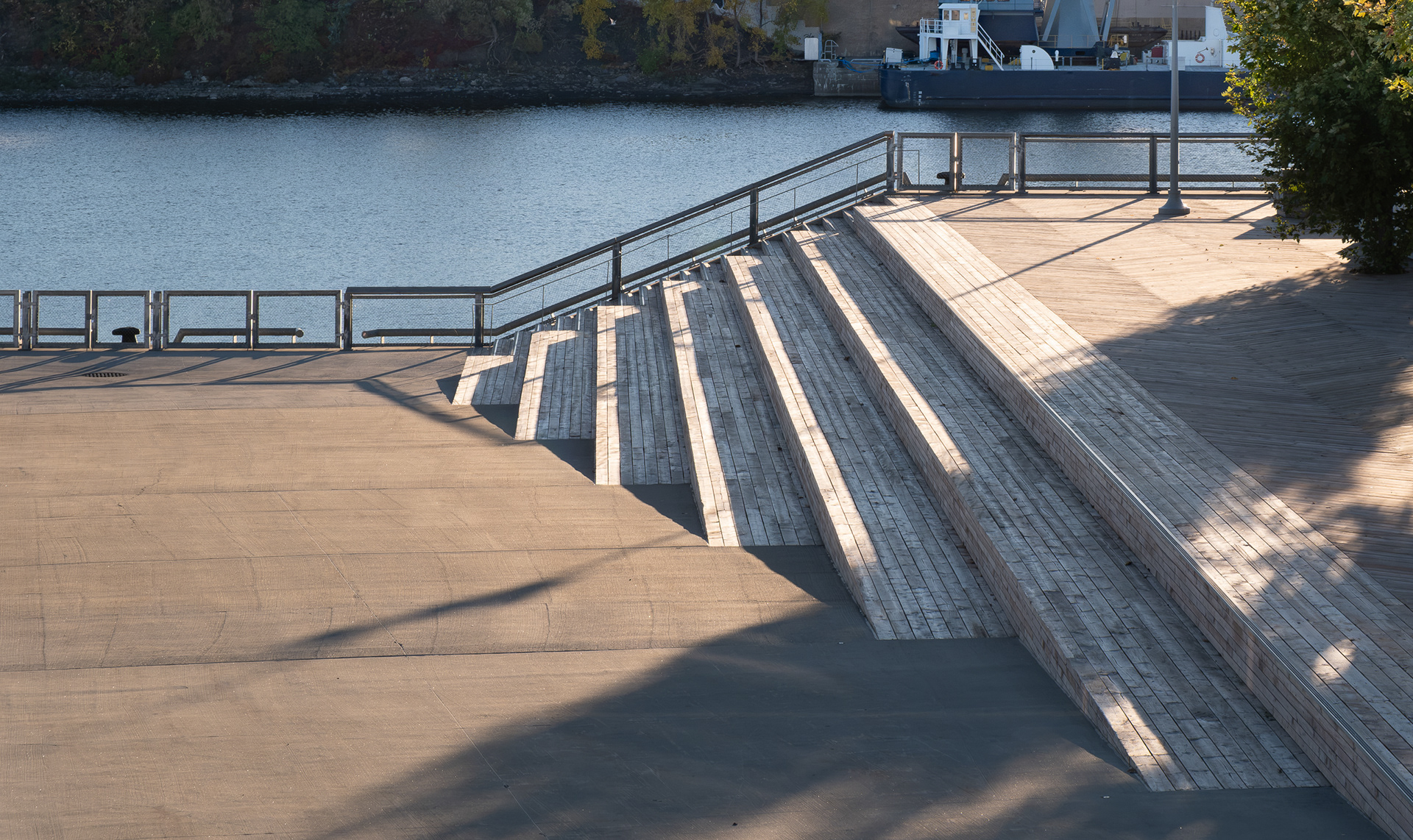 Gros plan sur les gradins en bois intégrés à l'espace public du Grand Quai de Montréal, conçus par NIPPAYSAGE et Provencher_Roy. L'ouvrage de transition en bois naturel descend vers une esplanade en béton près d’un garde-corps métallique, le tout bordant le fleuve Saint-Laurent en lumière naturelle de début de journée.
