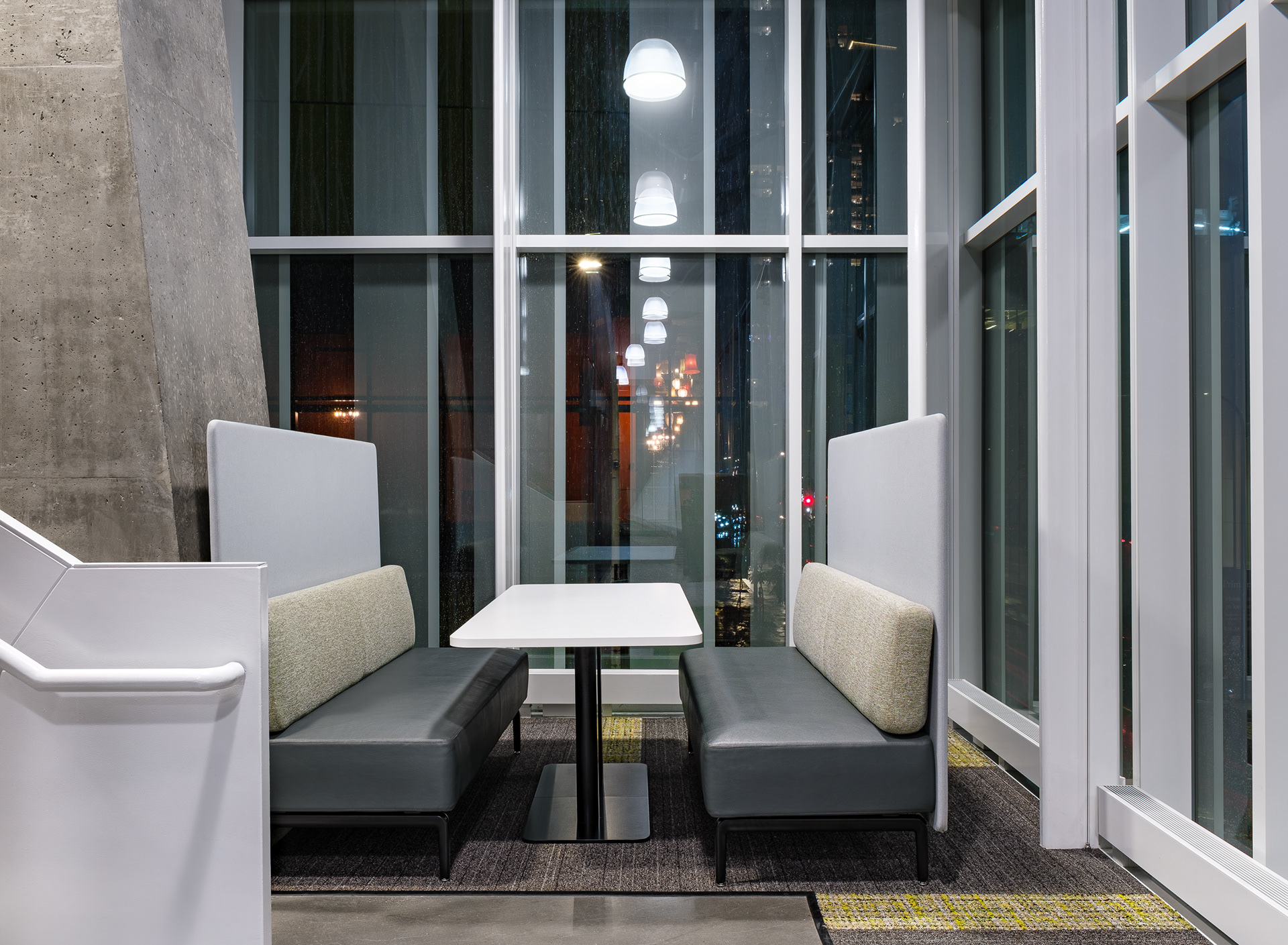 Zone de détente et de rencontre dans les espaces commerciaux de l'Îlot Balmoral. Deux banquettes modernes encadrent une table blanche contre la fenestration du mur-rideau, à côté d'une imposante structure en béton.