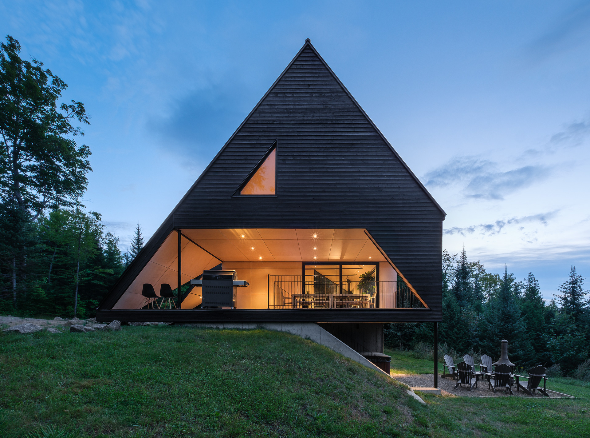 Vue latérale de la Cabine-A à Charlevoix par Bourgeois / Lechasseur au crépuscule. L'architecture triangulaire s'intègre au terrain en pente et révèle une grande terrasse couverte en bois clair ouverte sur la nature environnante.