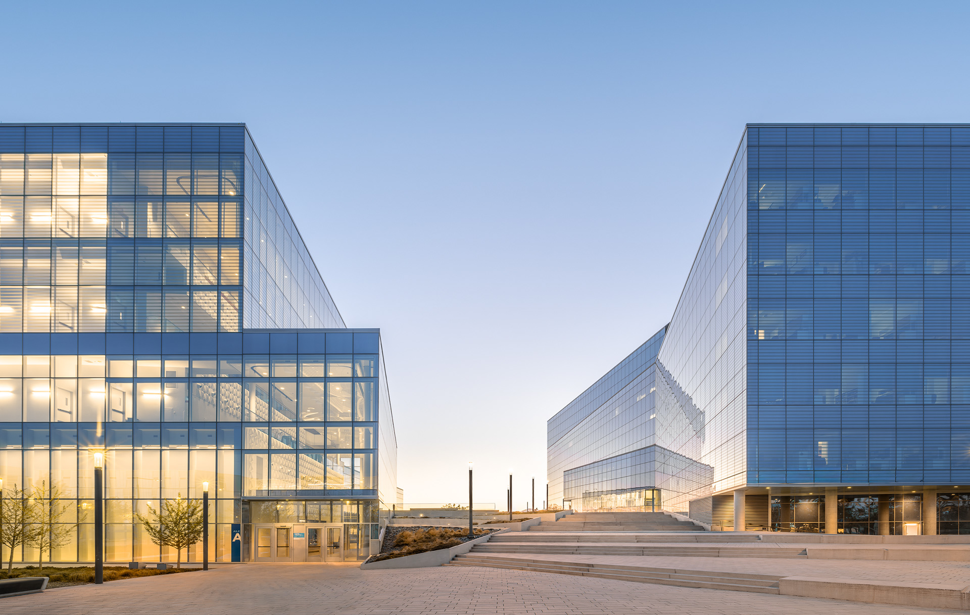 Perspective entre les pavillons du Complexe des sciences au Campus MIL de Montréal. Les larges emmarchements en béton et le calepinage guident le regard vers les façades lumineuses en verre fritté sous un ciel de fin de journée.