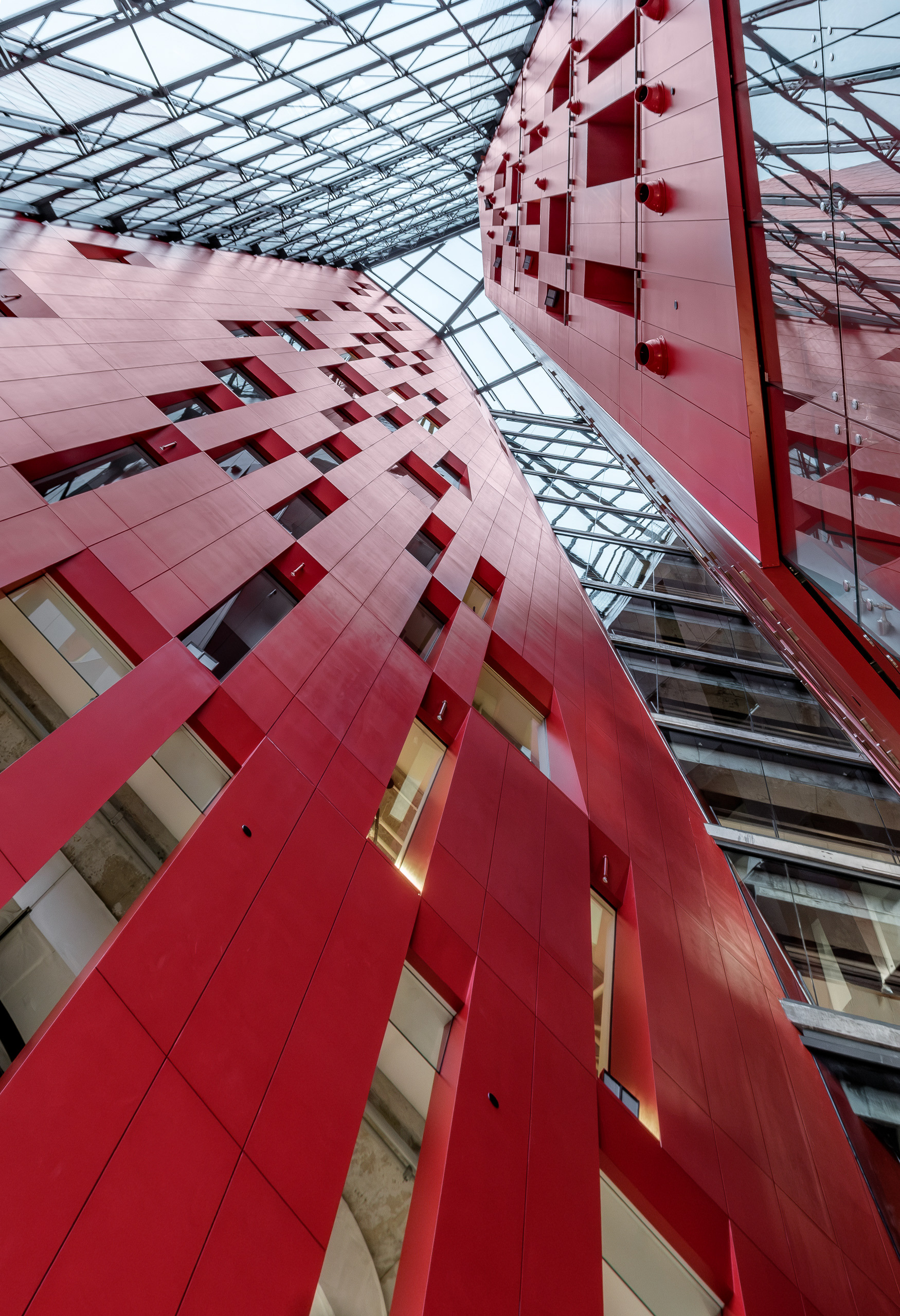 Perspective verticale dans l'atrium pleine hauteur de l'Îlot Balmoral à Montréal. Les monumentales façades intérieures rouges, percées de fenêtres géométriques, convergent vers une grande verrière structurelle.