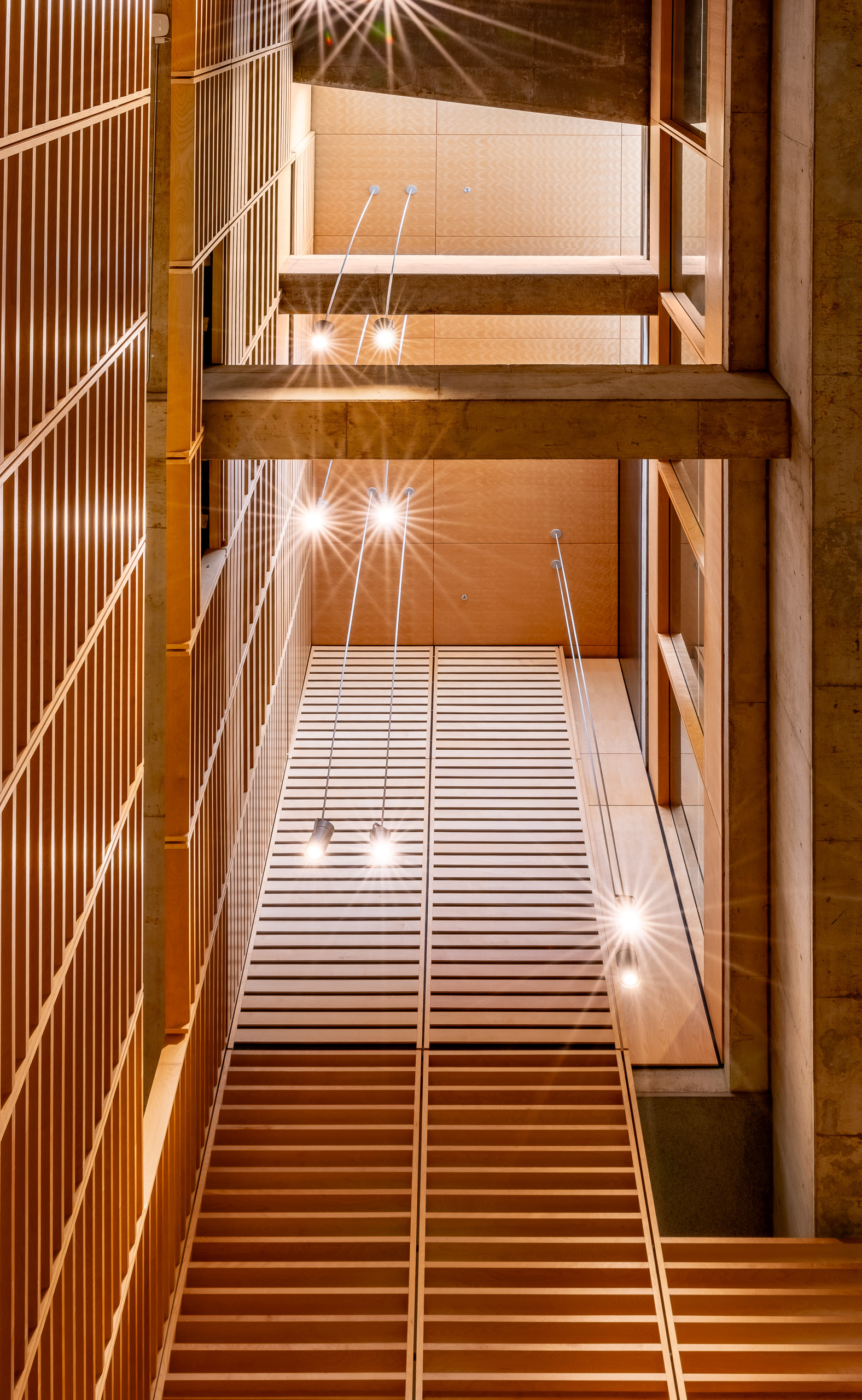 Vue en contre-plongée d'un puits de lumière à la Grande Bibliothèque de Montréal. L'architecture met en valeur un plafond et des murs revêtus de lattes de bois clair, traversés par des poutres en béton brut et des luminaires suspendus.