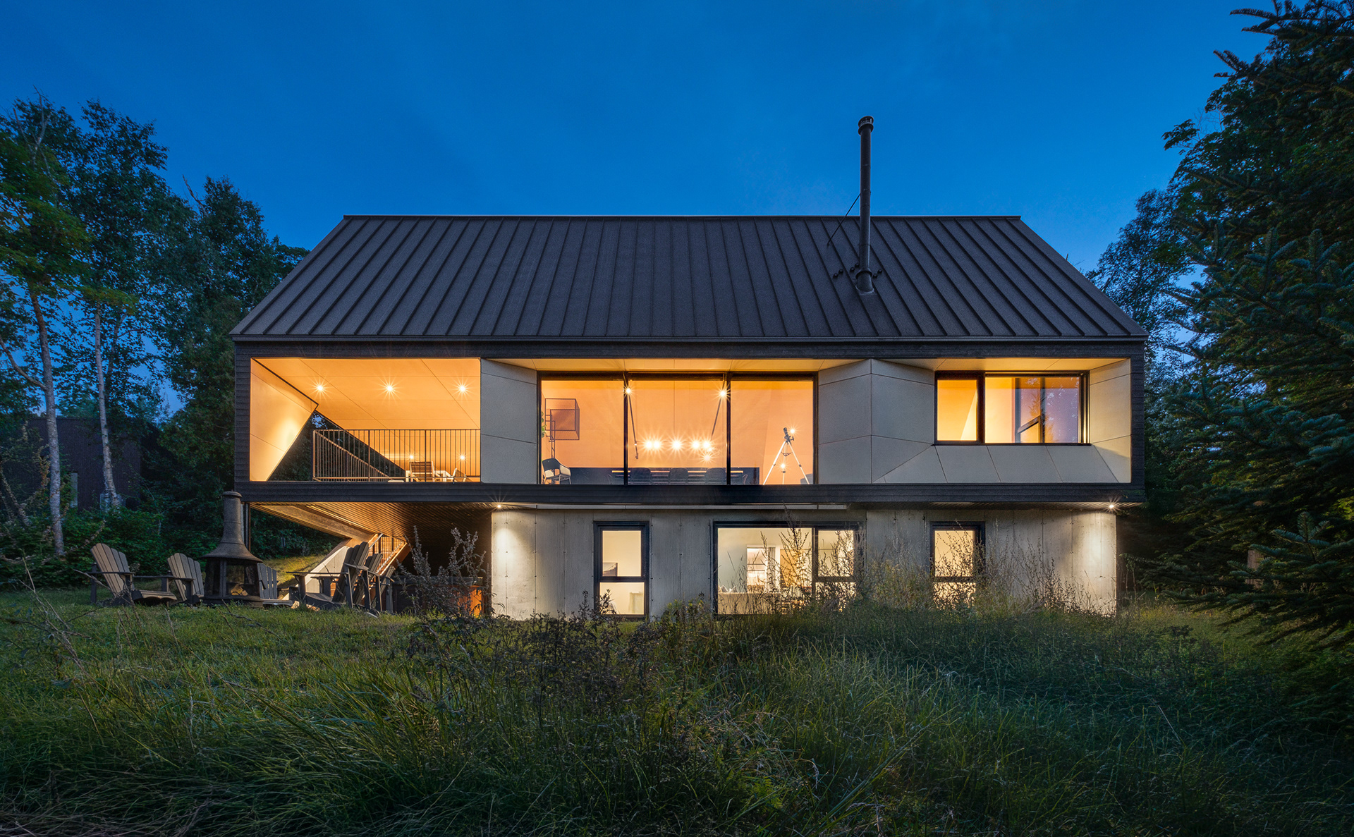 Vue de face de la résidence contemporaine Cabine-A à Charlevoix par Bourgeois / Lechasseur au crépuscule. L'architecture met en valeur un sous-sol en béton, une enveloppe en bois et une toiture à joint debout ancrés dans le paysage naturel.