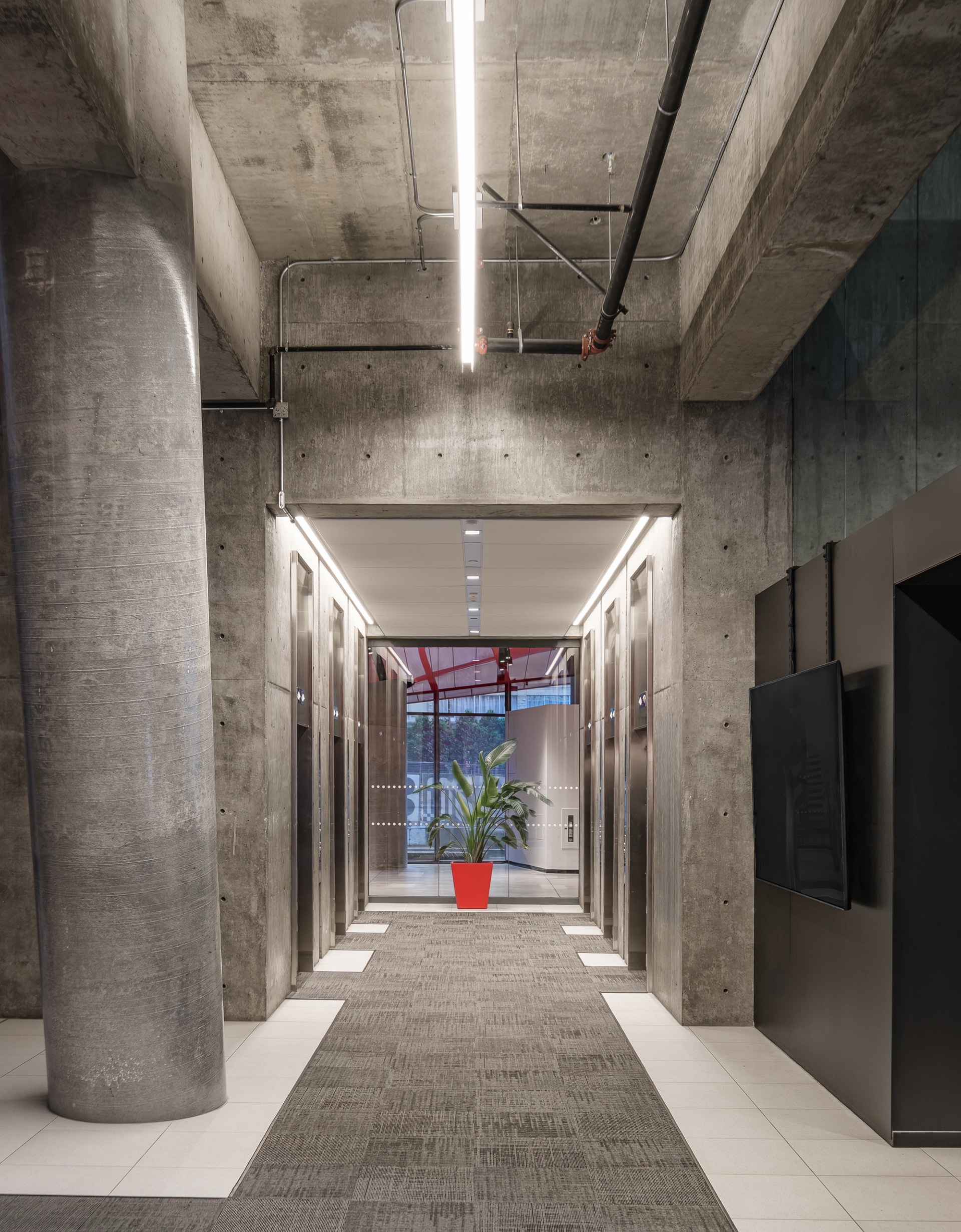 Hall des ascenseurs de l'Îlot Balmoral à Montréal. Cet espace de circulation affirme une esthétique industrielle avec son plafond et ses murs de béton brut, contrastant avec la vue fuyante vers le rouge éclatant de l'atrium.