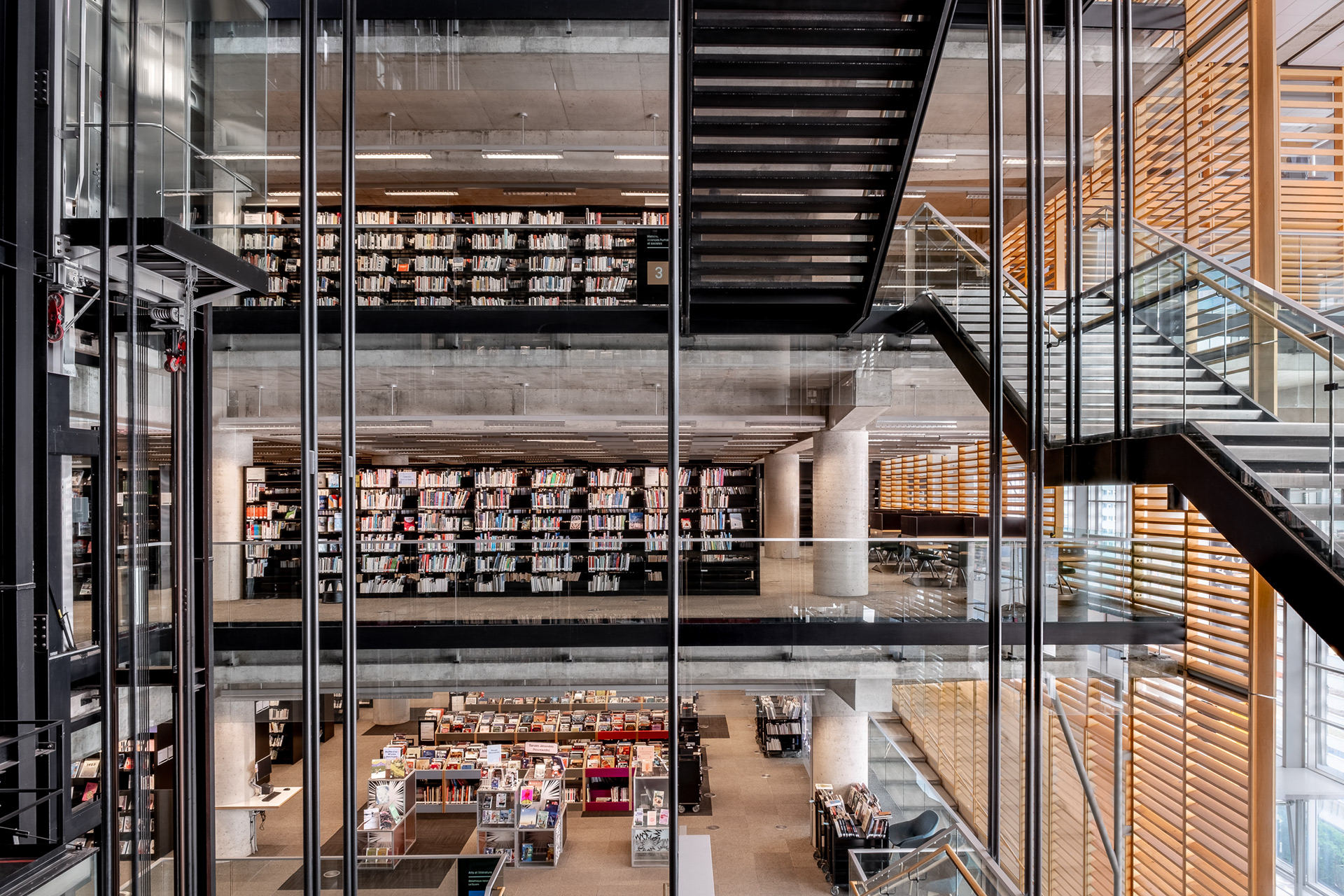 Perspective sur les différents étages de la Grande Bibliothèque de Montréal. À travers une paroi vitrée et des suspentes, on aperçoit les escaliers, les rayonnages de livres et la zone d'accueil au rez-de-chaussée.