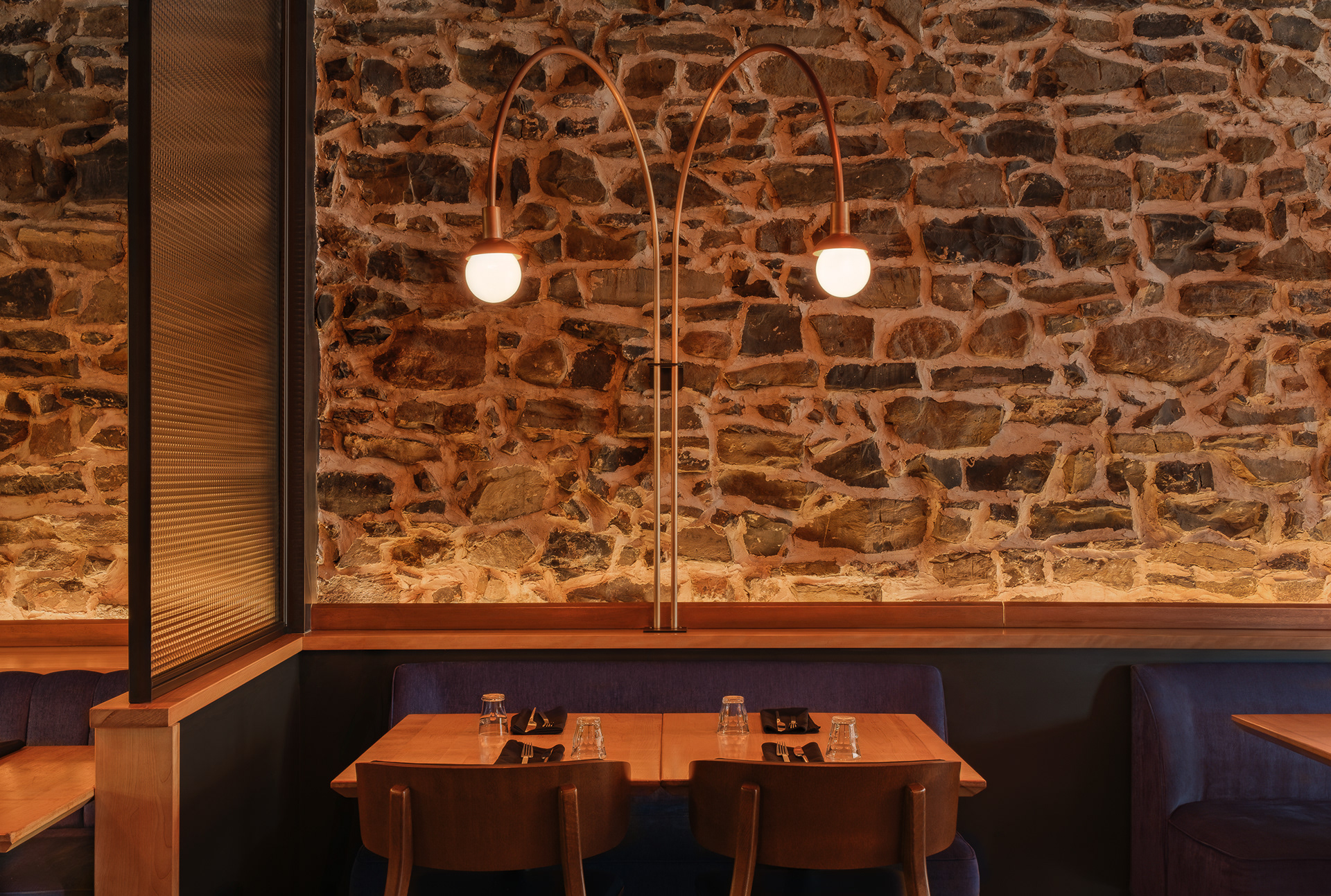 Détail de la salle à manger du restaurant Kwizinn à Montréal par Taktik Design. L'espace intègre une banquette bleue, une cloison en verre texturé et un luminaire sur mesure en cuivre devant un mur en maçonnerie patrimoniale.