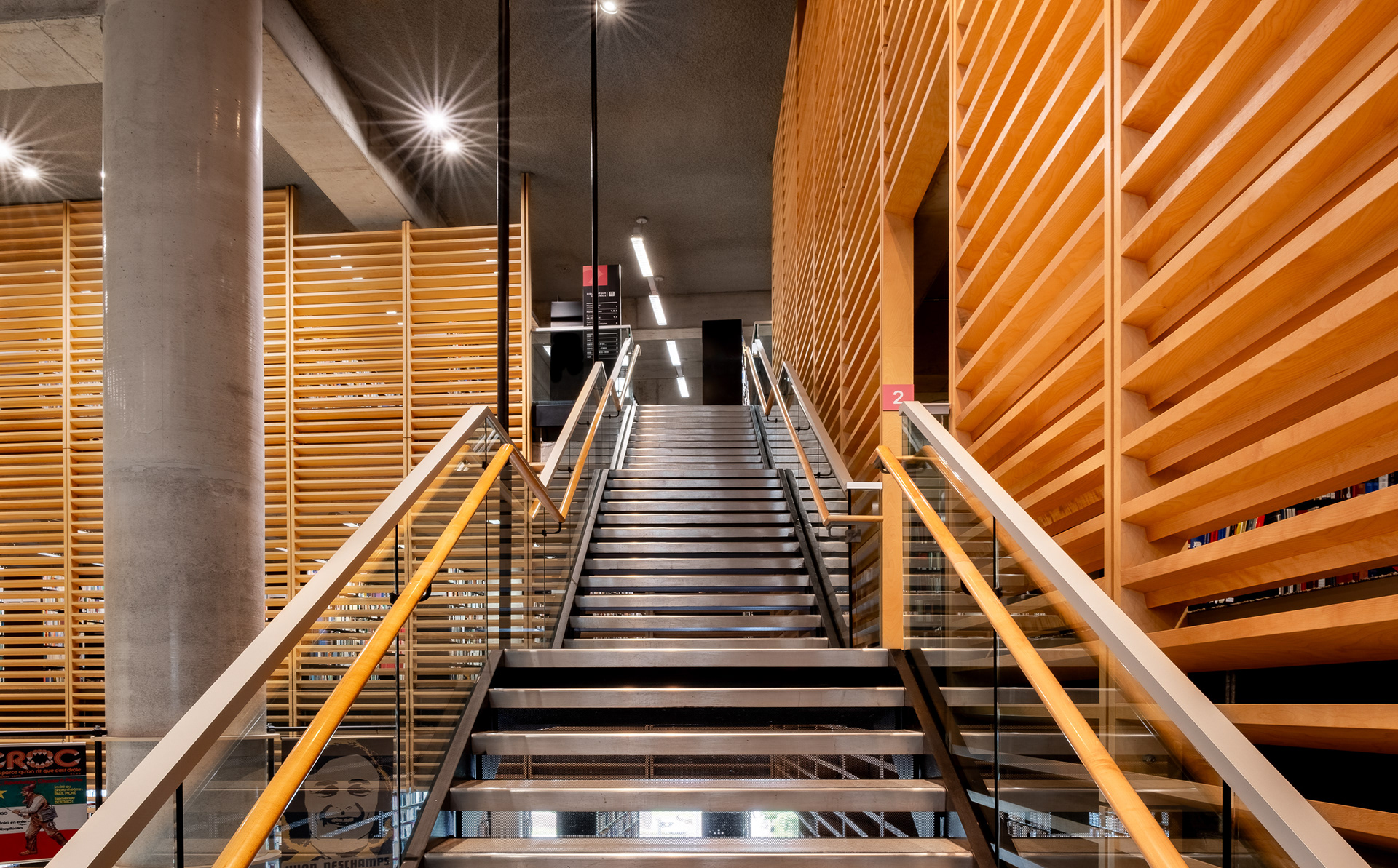 Vue frontale d'un escalier principal de la Grande Bibliothèque de Montréal. L'escalier en métal aux garde-corps vitrés monte à travers la structure, encadré par des parois monumentales en claires-voies de bois et un plafond en béton.