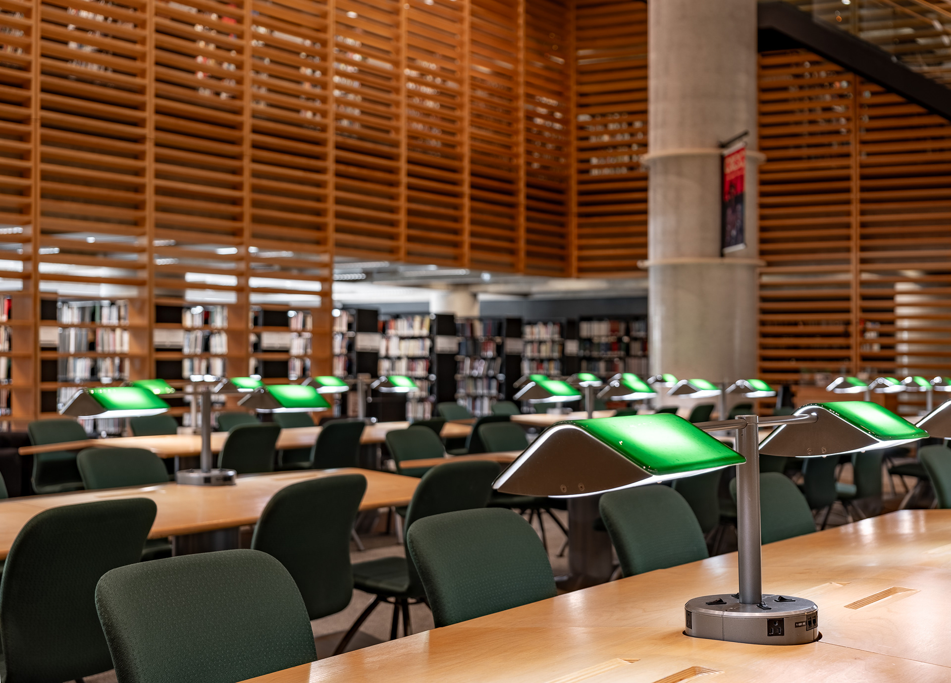 Perspective sur l'espace de consultation de la Grande Bibliothèque de Montréal. De longues tables de travail en bois et leurs lampes vertes caractéristiques s'alignent devant un haut mur en claires-voies de bois et des étagères de livres.
