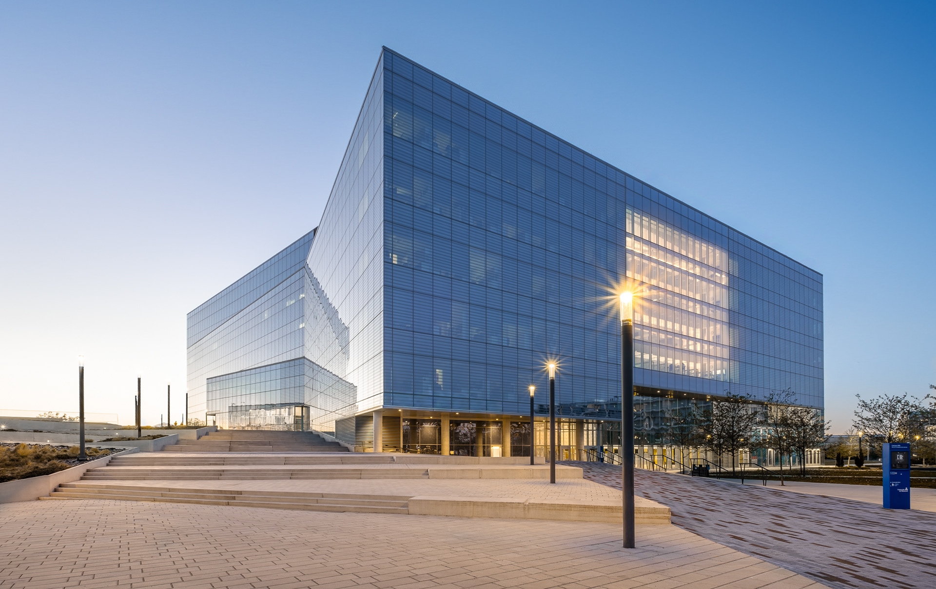 Vue d'ensemble du Complexe des sciences du Campus MIL à Montréal au crépuscule. L'imposante façade en verre fritté s'élève au-dessus d'une vaste place publique caractérisée par de larges emmarchements en béton et un calepinage texturé.