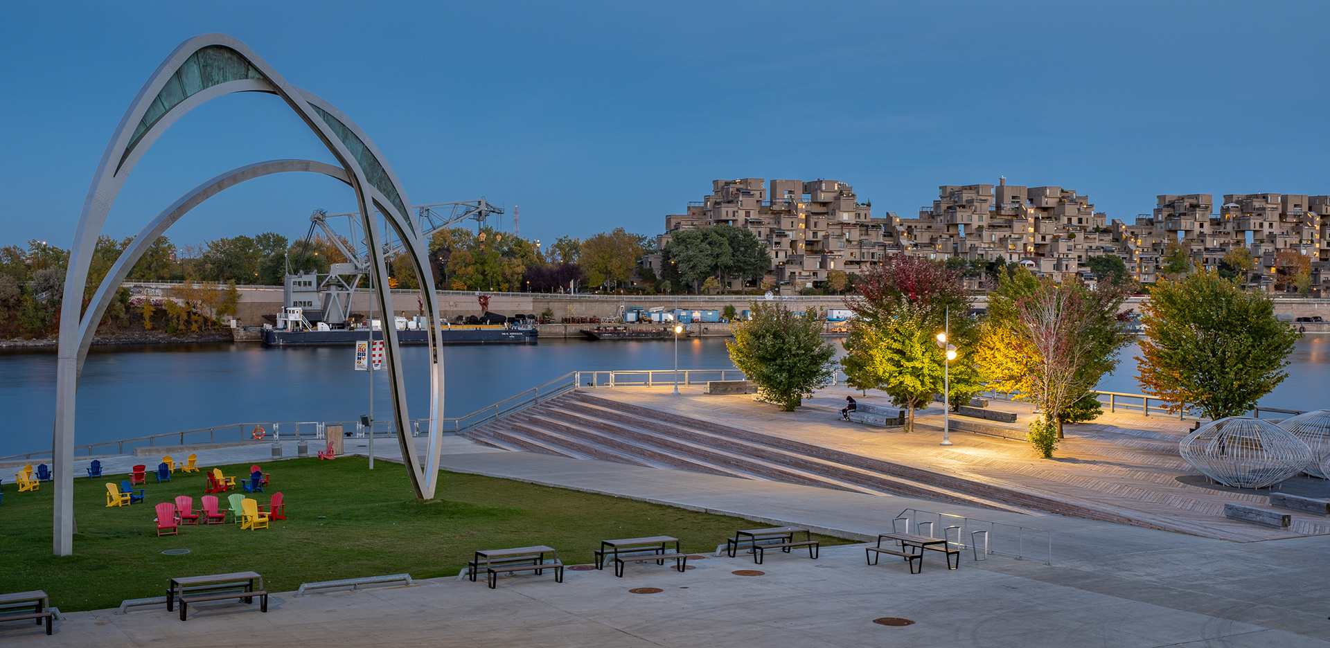 Vue d’ensemble du Grand Quai du Port de Montréal au crépuscule, conçu par NIPPAYSAGE et Provencher_Roy. On y voit l’œuvre d'art public de Yann Pocreau à gauche, un vaste espace piétonnier avec des gradins et un platelage en bois, du mobilier urbain coloré, ainsi que l'ensemble résidentiel Habitat 67 en arrière-plan au-delà du fleuve.