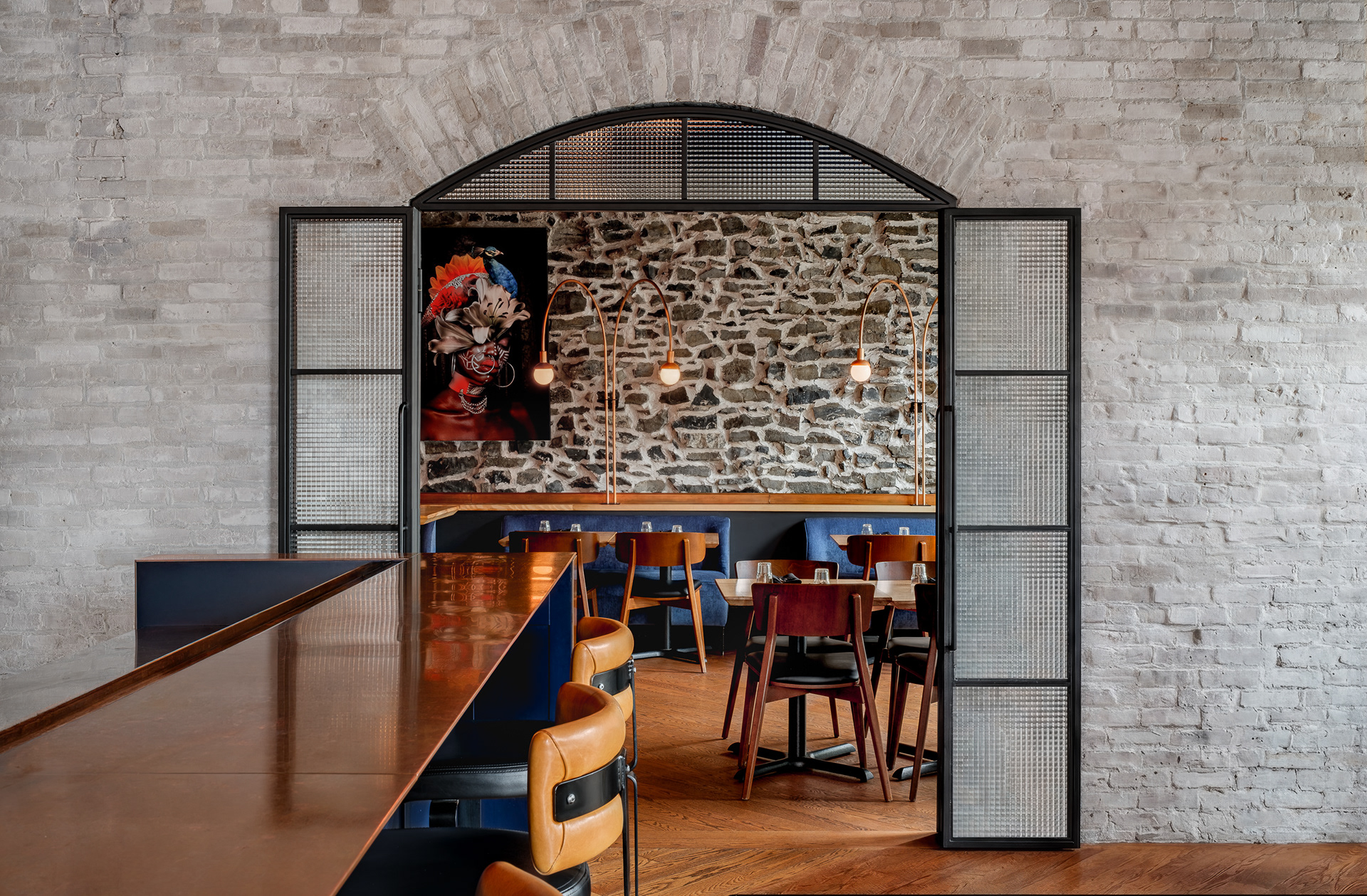 Portes en verre texturé ouvertes révélant une salle à manger intime au restaurant Kwizinn, caractérisée par un mur en pierre brute, une banquette bleue, des éclairages sur mesure en cuivre et une toile colorée.