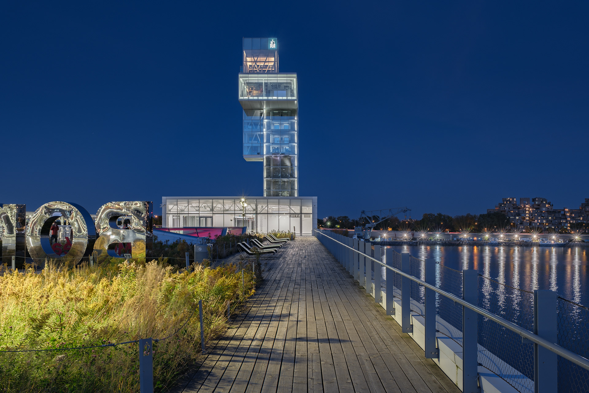 Vue nocturne de la tour d’observation illuminée du Grand Quai de Montréal, par NIPPAYSAGE et Provencher_Roy. La passerelle en bois, bordée d'une toiture végétalisée et d'une œuvre en acier, s'étire le long du fleuve Saint-Laurent.