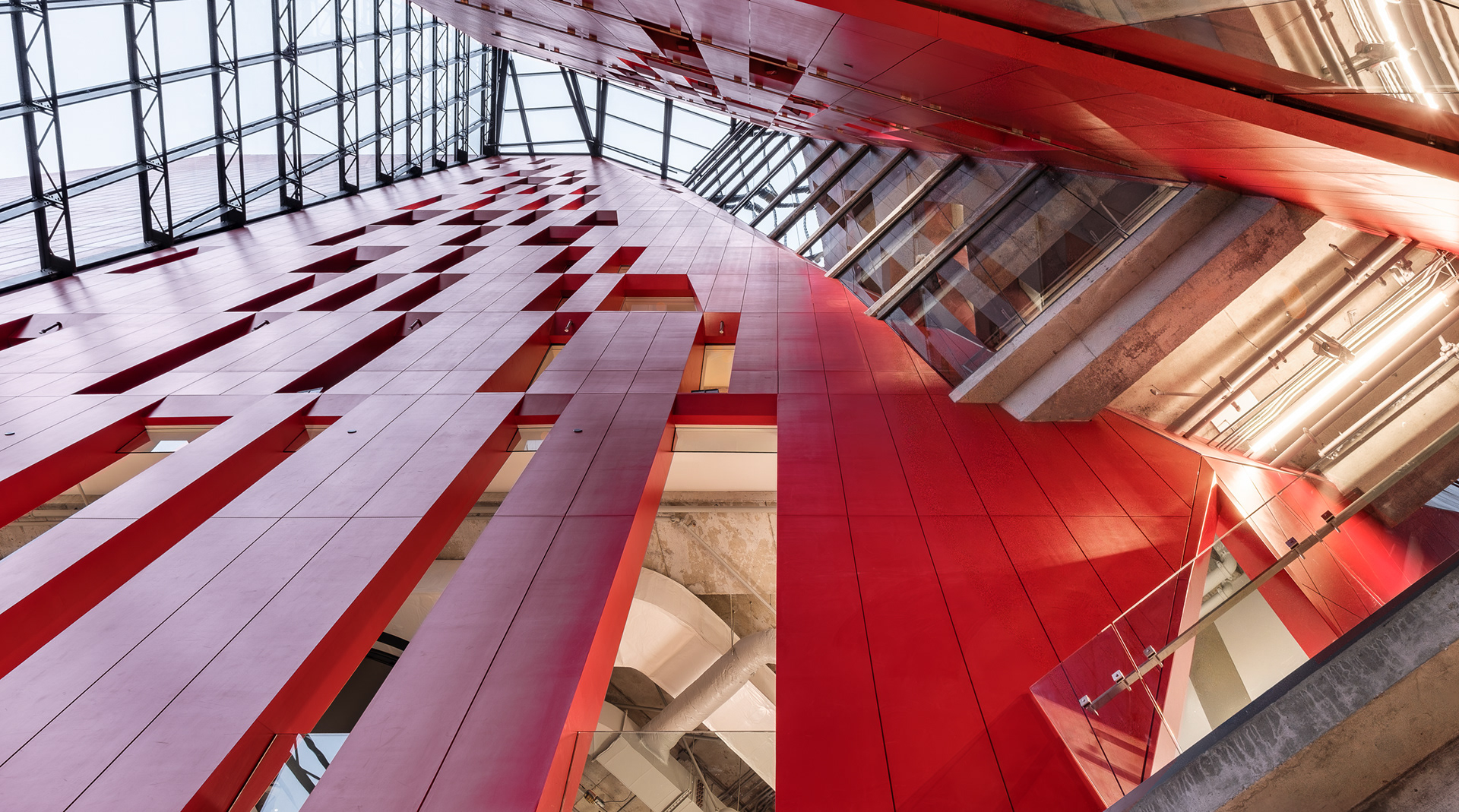 Vue en contre-plongée de l'atrium de l'Îlot Balmoral conçu par Provencher_Roy. La structure intérieure expose des parois rouges angulaires, des éléments en béton et le dessous des passerelles suspendues sous la verrière.