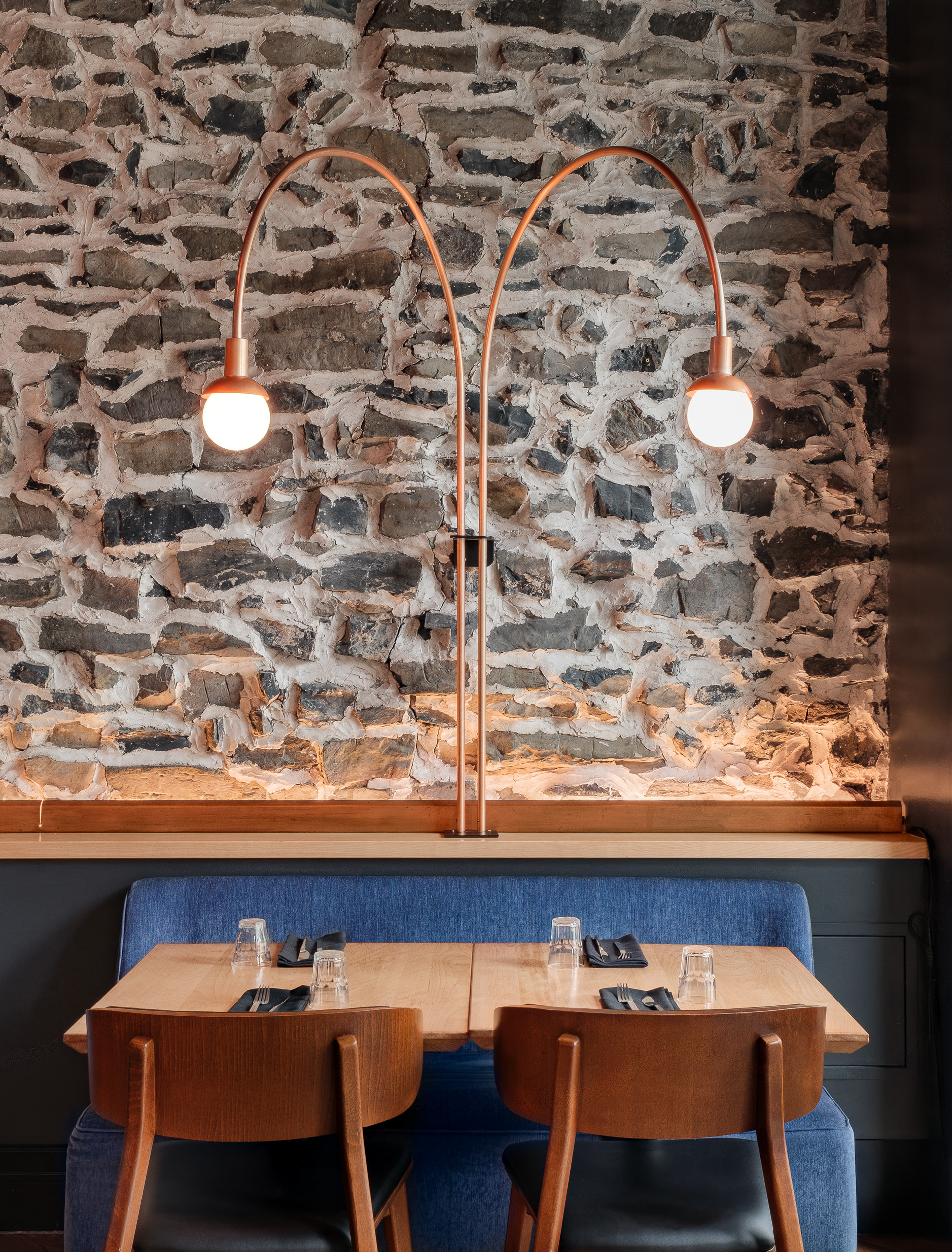 Gros plan sur l'aménagement intérieur du restaurant Kwizinn par Taktik Design à Montréal. Une table en bois et une banquette bleue sont mises en valeur par un luminaire sur mesure en cuivre à double arc adossé à un mur en maçonnerie brute.
