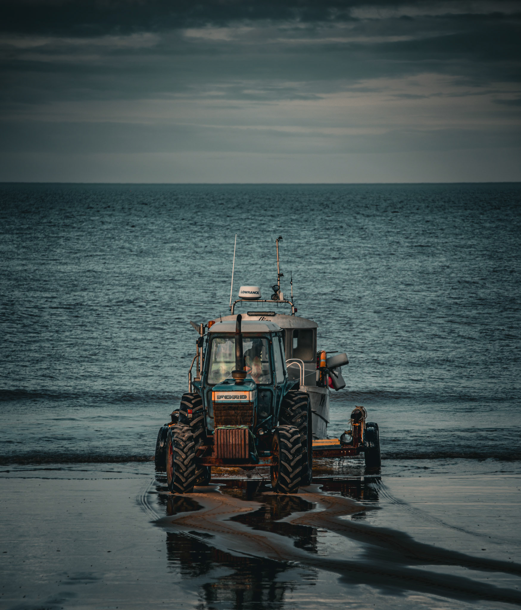 Against the backdrop of the vast ocean, the sturdy 'Ford' tractor diligently tends to its duties, leaving imprints of hard work and determination on the sandy shores.