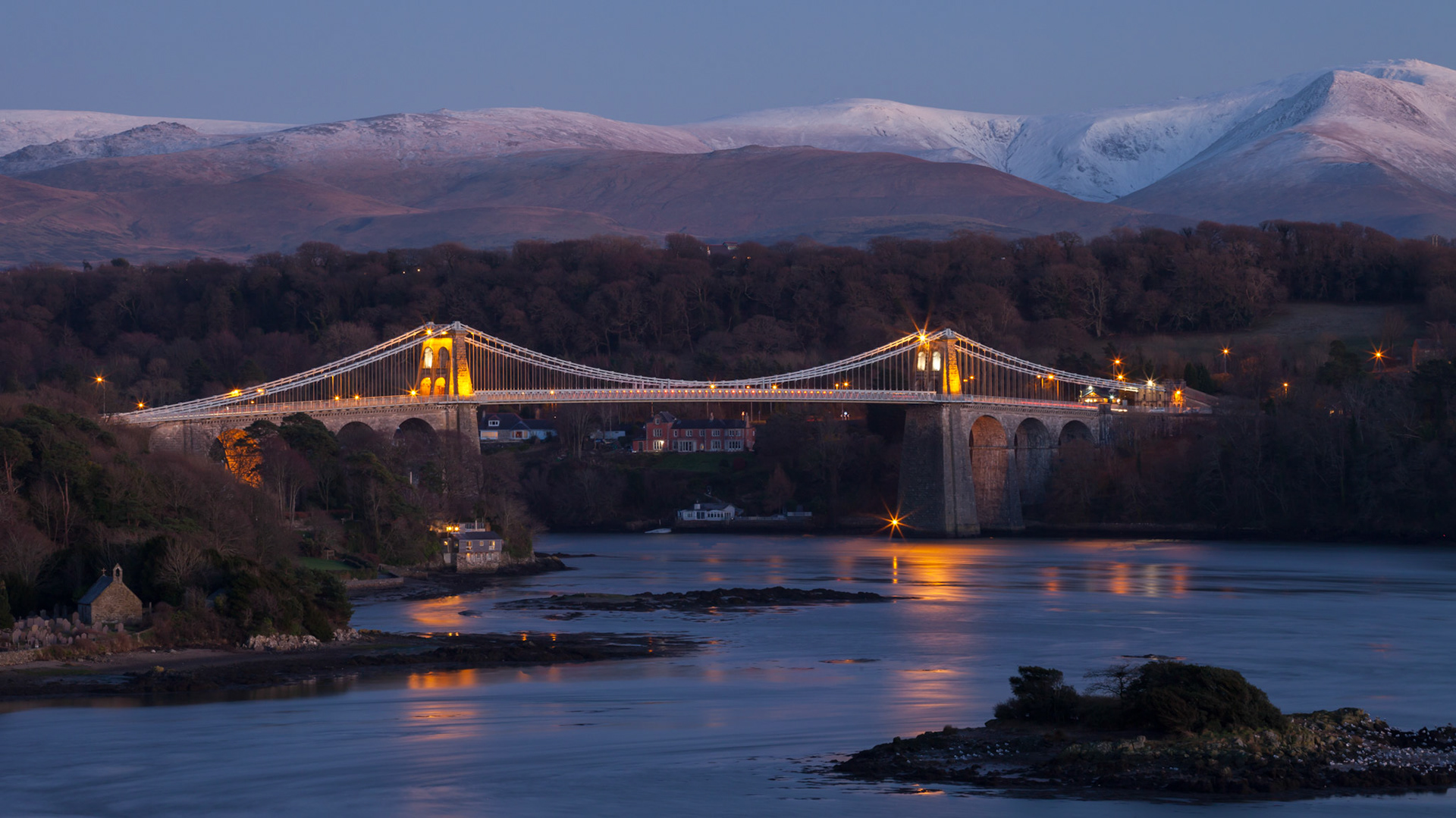 Menai Bridge at dusk with snow capped mountains in the background and lights on the bridge, winter, Anglesey, Wales, UK
