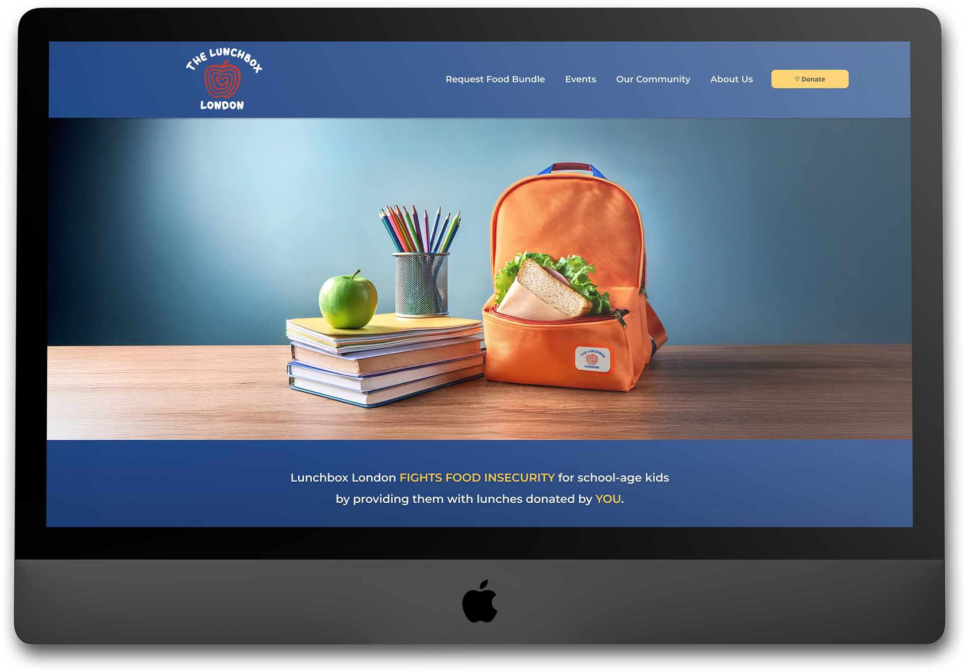 A computer monitor displays the full web version of the redesigned Lunchbox London landing page. The hero image shows a bright lunchbag, flanked by school books and an apple. The dominant color is blue.
