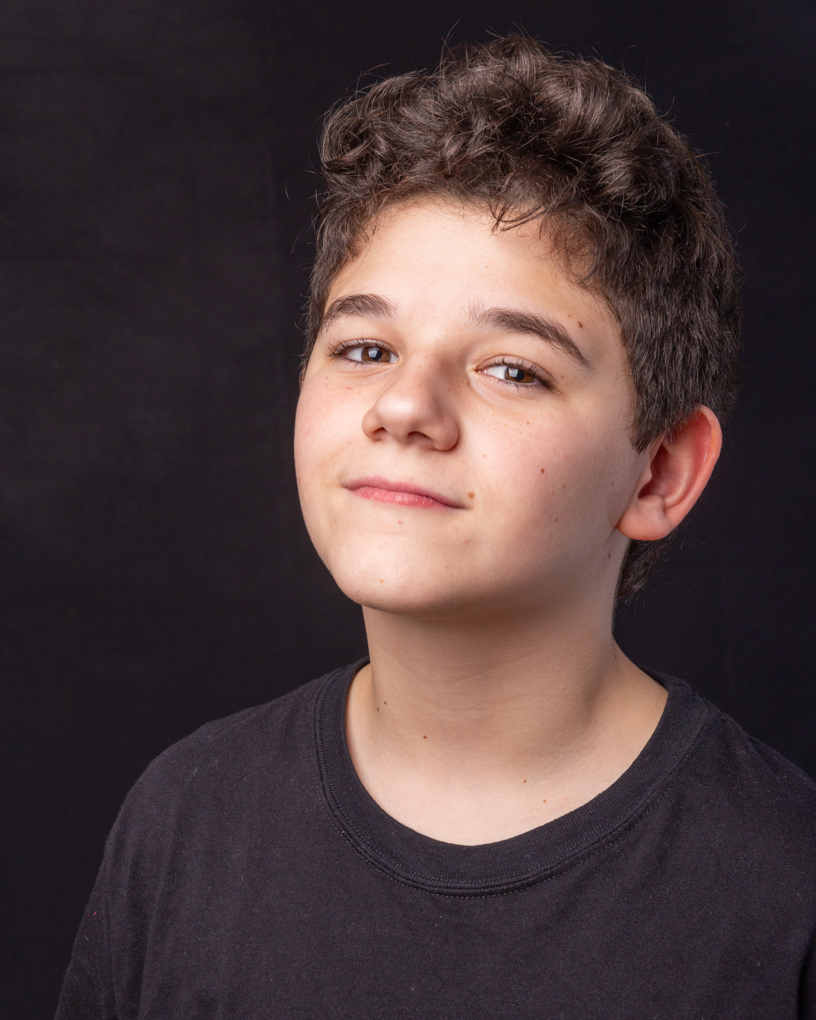 Retrato de primer plano de un adolescente de pelo castaño rizado sobre un fondo negro de estudio, mirando a la cámara con una sonrisa sutil y segura. Fotografía por Pau Aiello.