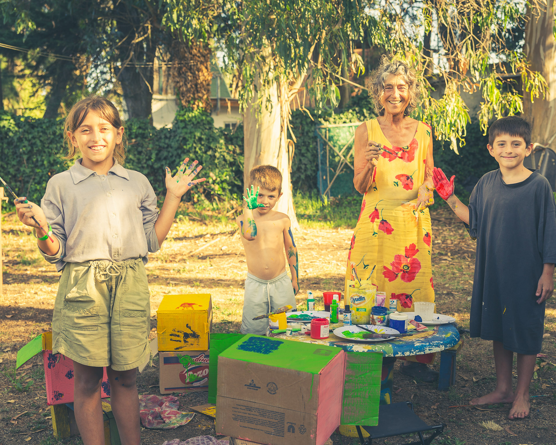 Una abuela y sus tres nietos se divierten pintando cajas de cartón al aire libre, con las manos manchadas de pintura, en una sesión de fotos documental familiar.