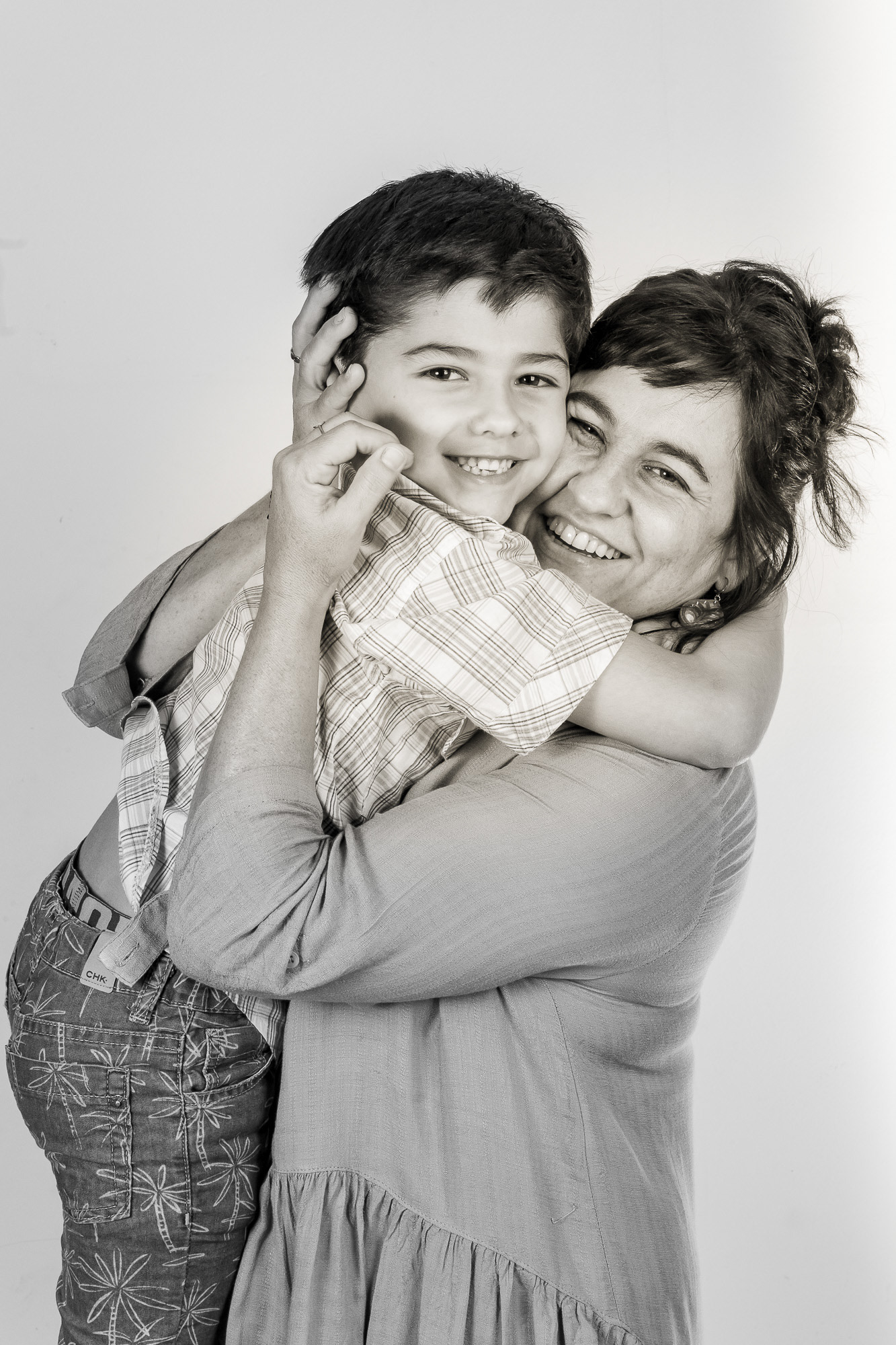 Retrato en blanco y negro de una madre abrazando a su hijo pequeño, ambos sonriendo con genuina alegría y afecto en un estudio. Fotografía de Pau Aiello.