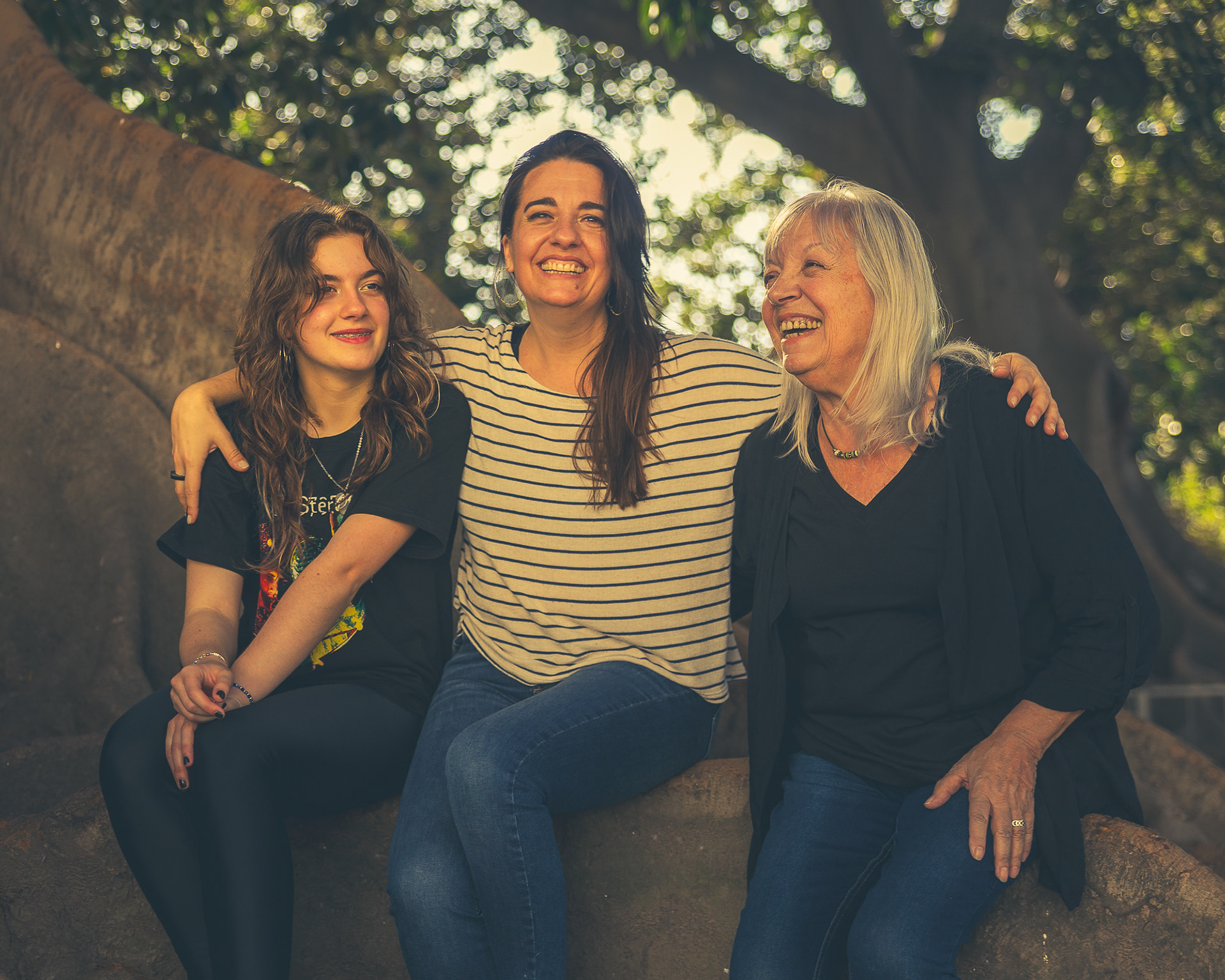 Tres generaciones de una familia —abuela, madre e hija— ríen juntas sentadas en la raíz de un árbol, en una sesión de fotos documental al aire libre por Pau Aiello.
