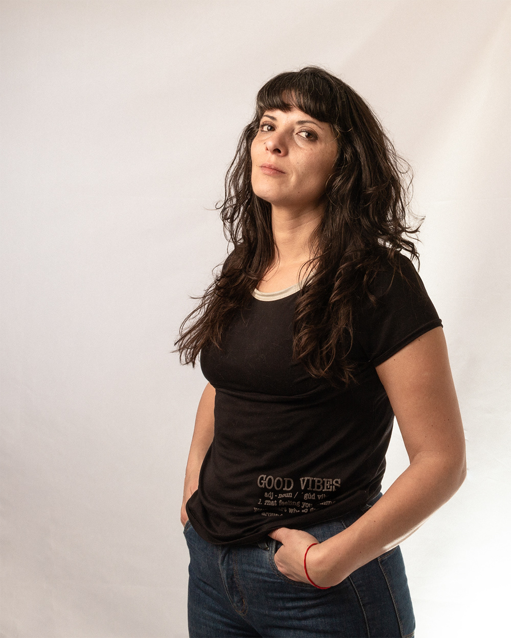 Autorretrato de Pau Aiello, fotógrafa, de pie sobre un fondo blanco luminoso. Viste un jean y una remera negra. Mira directamente a la cámara con una expresión serena y las manos en los bolsillos.
