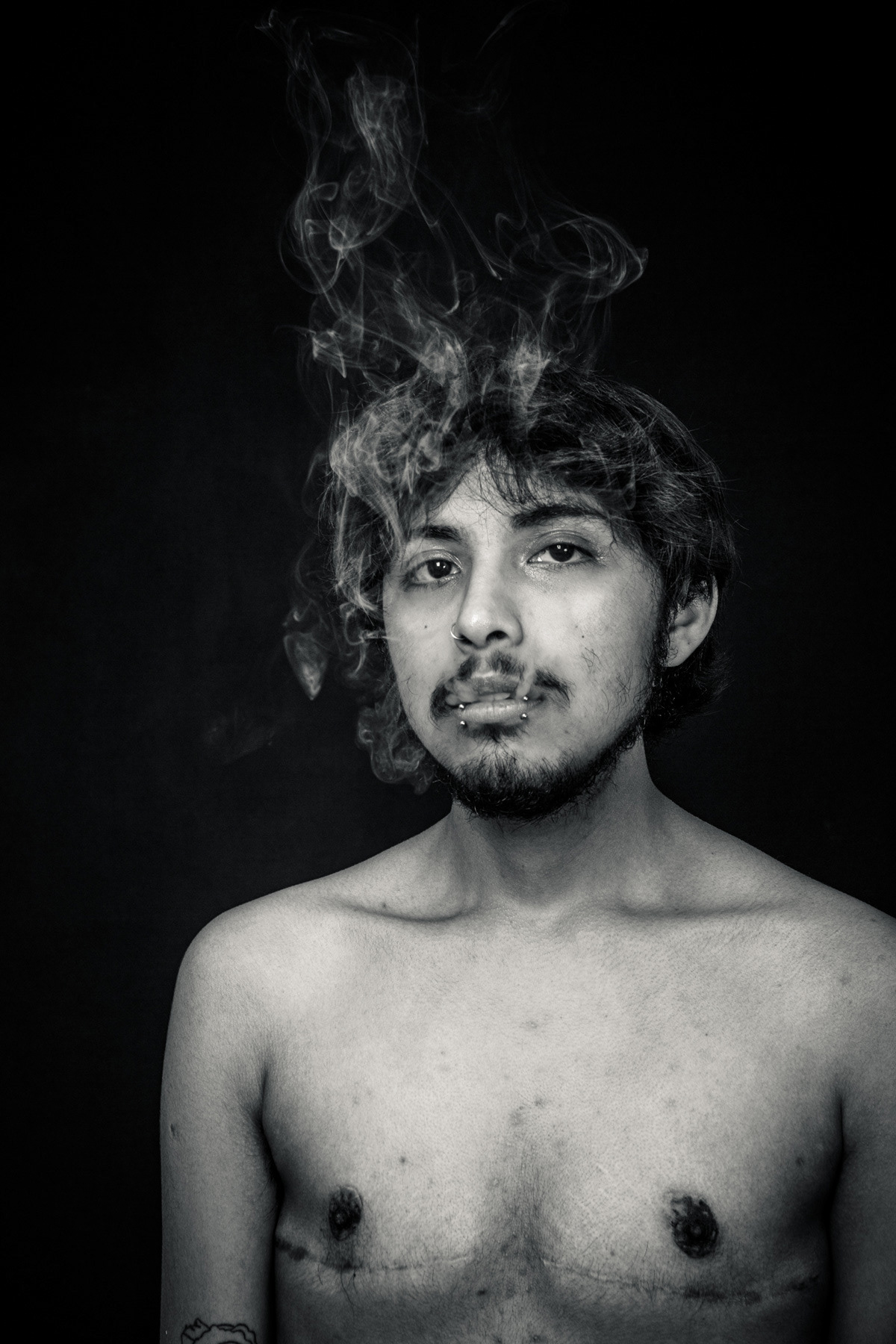 Retrato en blanco y negro de un actor trans joven en estudio, sobre fondo negro, largando humo hacia su rostro, fotografía de Pau Aiello en Buenos Aires.
