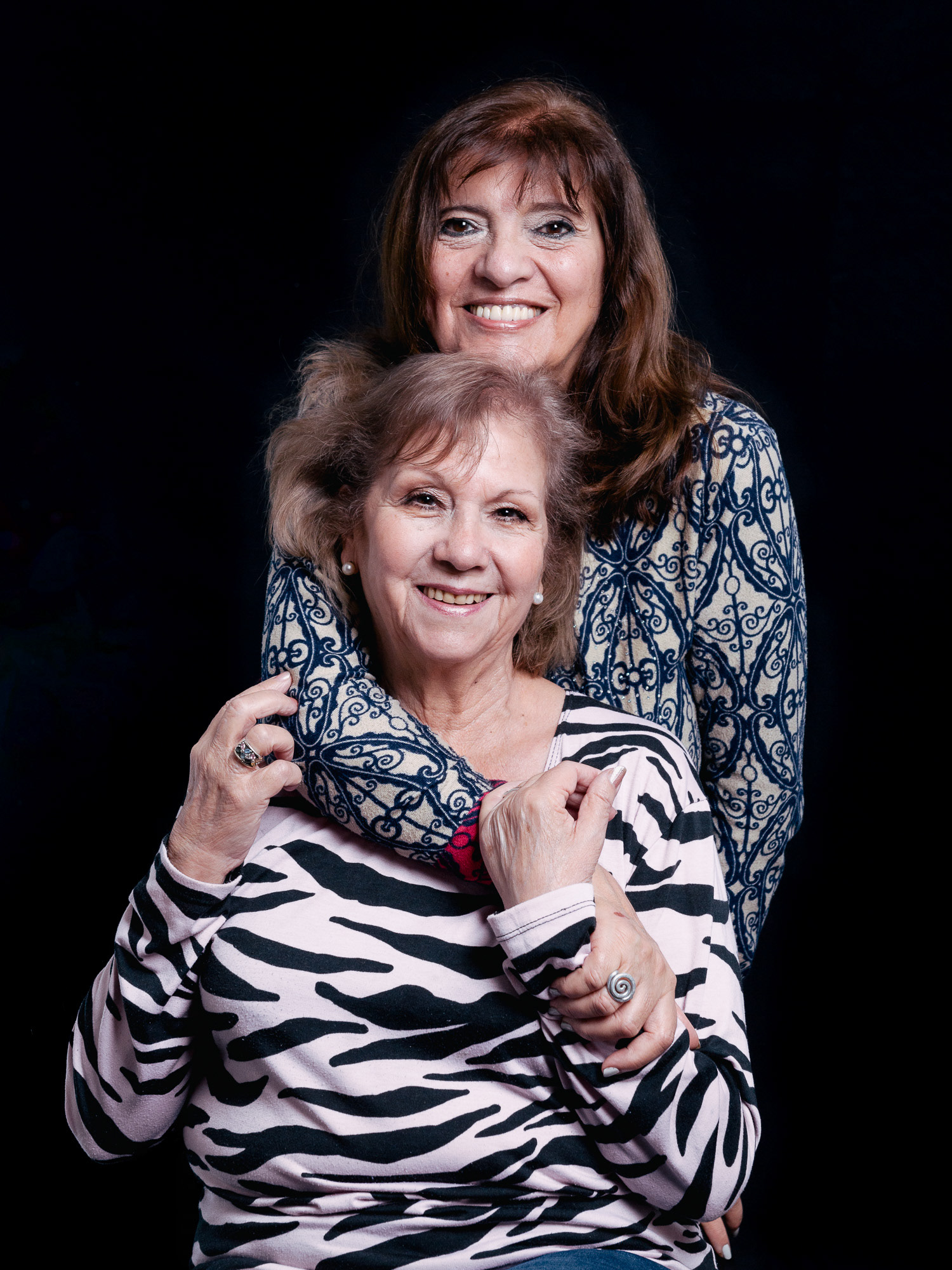 Retrato de estudio sobre fondo negro de dos hermanas adultas sonriendo, una abrazando a la otra por los hombros en un gesto de cariño. Fotografía por Pau Aiello.