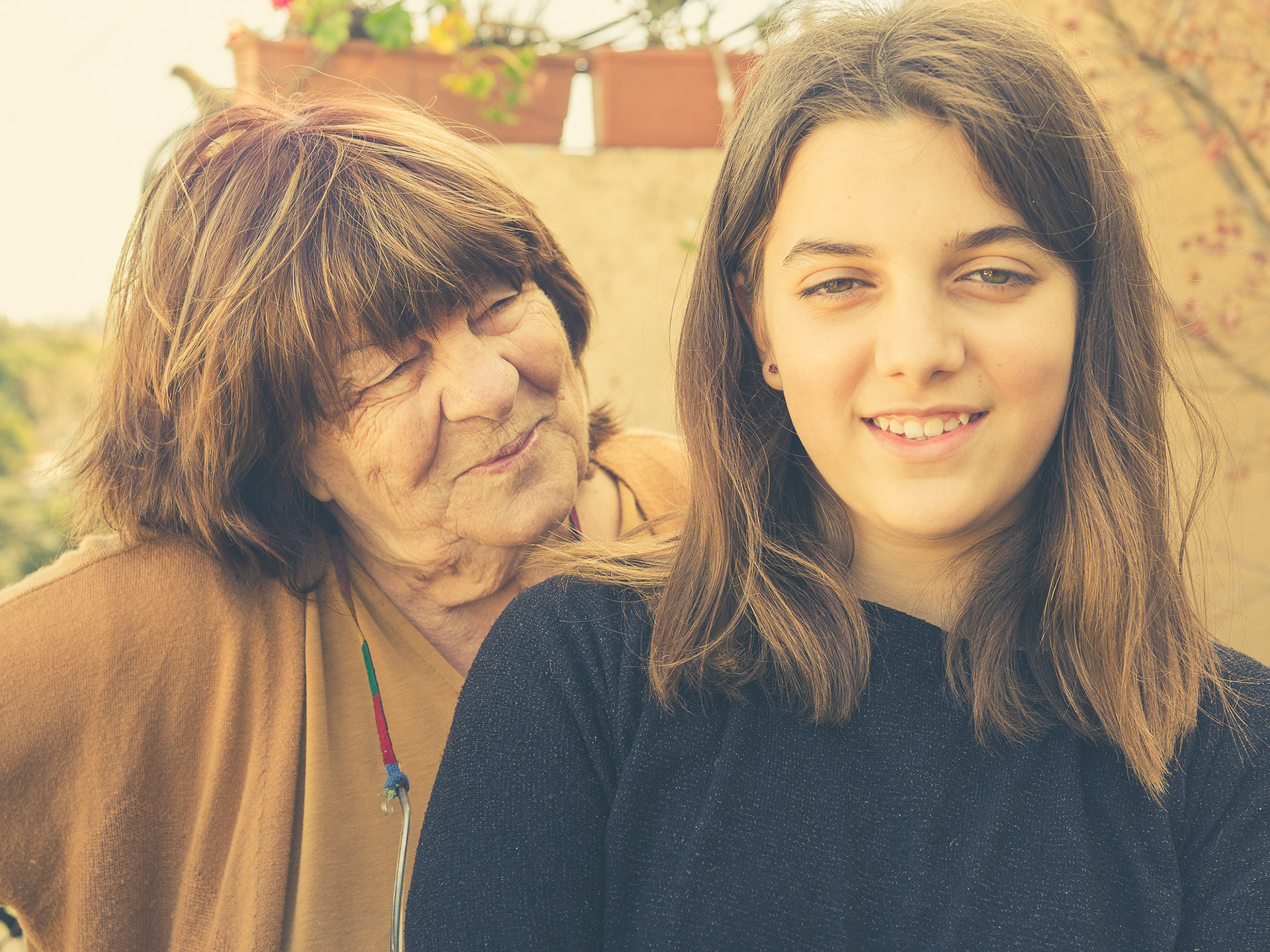 Primer plano de una nieta adolescente sonriendo a cámara, mientras su abuela la mira por detrás con una expresión de afecto y complicidad.