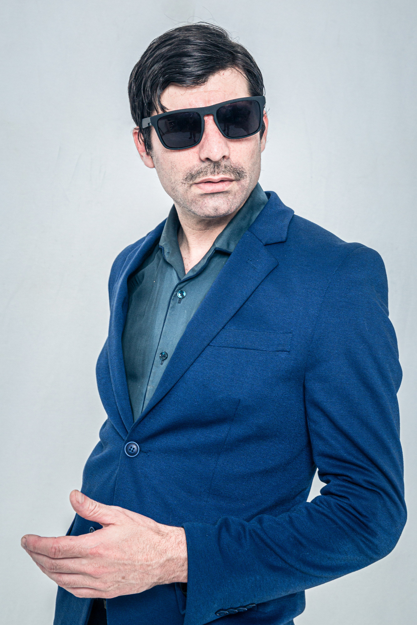 Retrato de estudio de un actor en personaje, vistiendo un traje azul y gafas de sol, con una expresión seria y un gesto corporal dinámico. Fotografía por Pau Aiello.