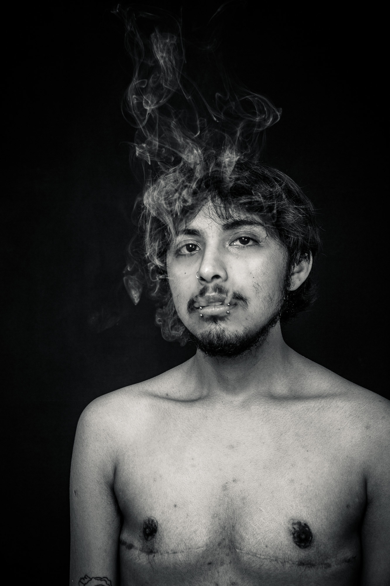 Retrato íntimo en blanco y negro de un hombre trans con cicatrices de mastectomía, mirando directamente a la cámara mientras humo se eleva desde su cabeza. Fotografía de Pau Aiello.