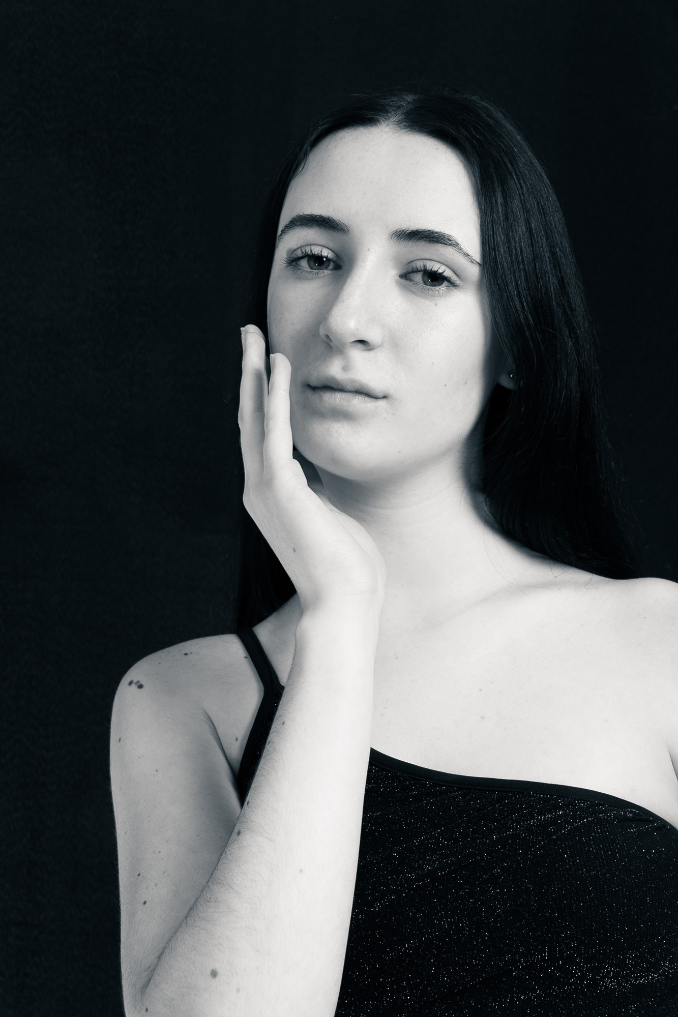 Retrato clásico en blanco y negro de una joven modelo con pelo largo y oscuro, posando con la mano en la mejilla sobre un fondo negro de estudio. Fotografía por Pau Aiello.