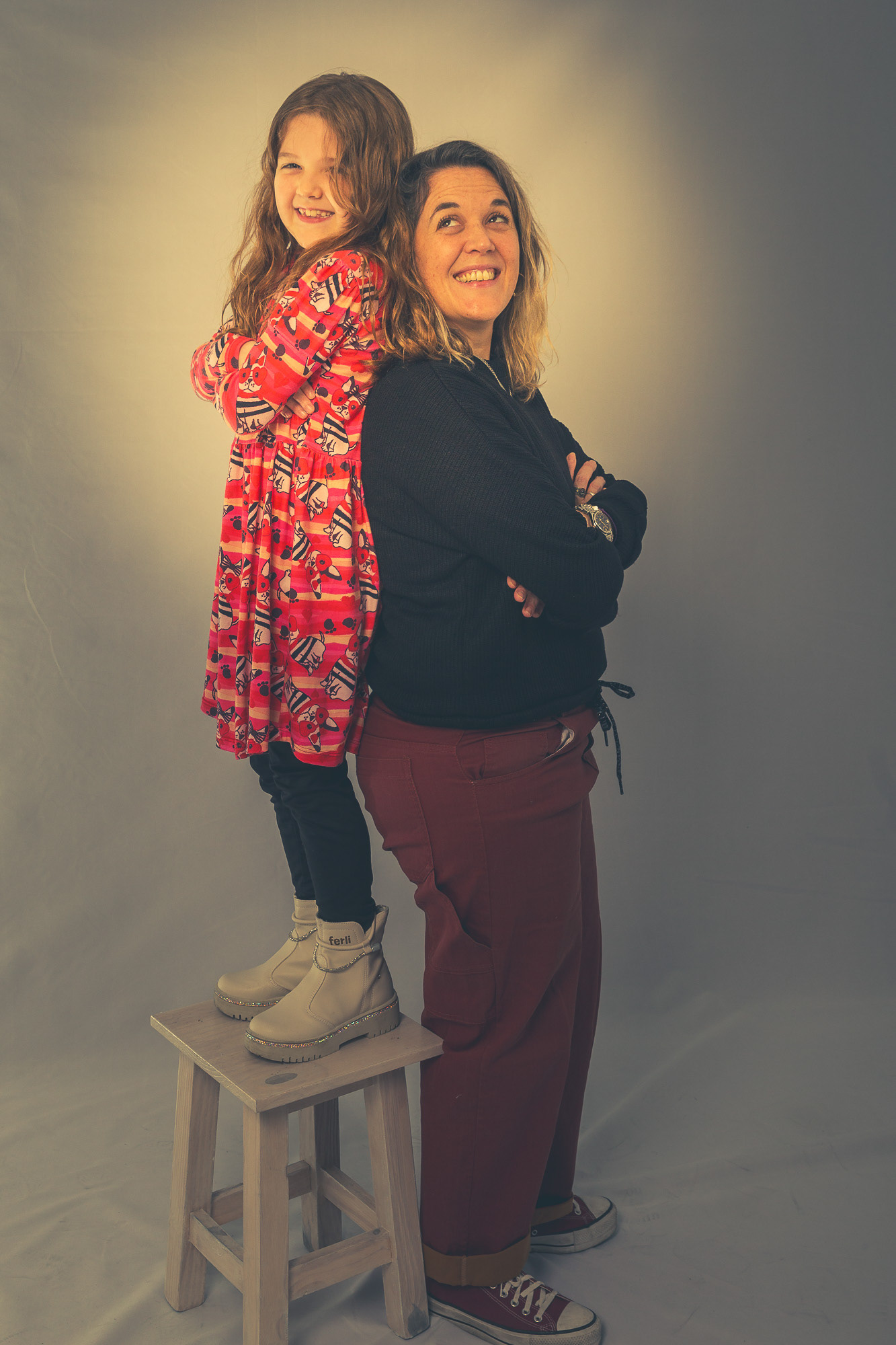 Madre e hija riendo en el estudio, dándose la espalda mientras la niña, parada en un banquito, queda más alta que su mamá.