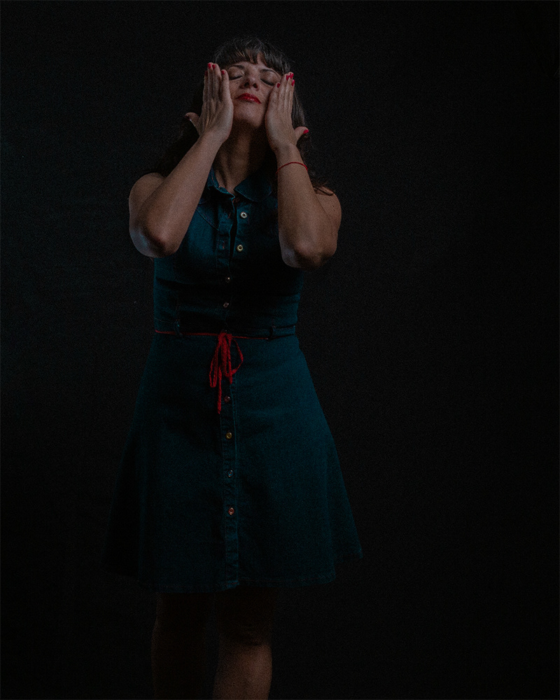 Autorretrato de Pau Aiello sobre un fondo negro. La luz es dramática y lateral. Tiene los ojos cerrados y se cubre parte del rostro con las manos en un gesto íntimo y reflexivo. Viste un vestido oscuro.