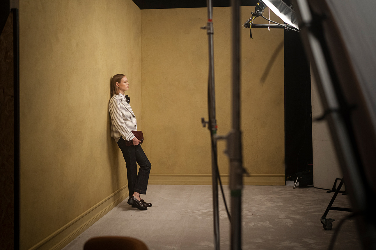 LUISA CERANO SS2026 Kampagne: Mode-Model in gestreiftem Blazer, weißem Hemd und schwarzen Hosen lehnt an Wand mit Beleuchtungsausrüstung im Studio