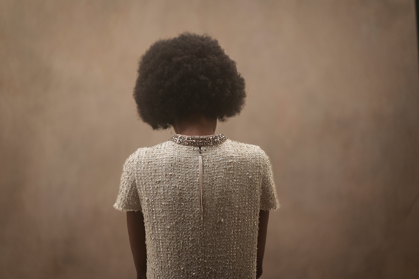 Rückansicht des Models mit Afro in funkelndem Tweed-Kleid mit Reißverschluss und Halskette vor beigem Hintergrund im Studio der LUISA CERANO Frühling/Sommer 2026