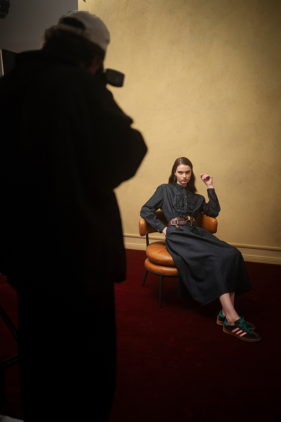 Fotograf erfasst Model in dunklem Denim-Kleid, Doppelgürtel und Sneakers auf Stuhl in schwach beleuchtetem Studio der LUISA CERANO Frühling/Sommer 2026
