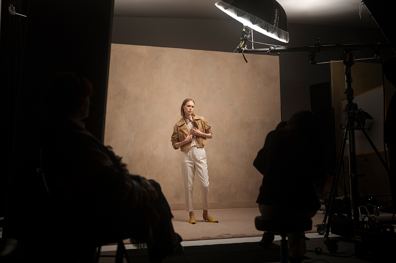 Gruppenaufnahme hinter den Kulissen der LUISA CERANO SS2026 Kampagne: Model in brauner Jacke, weißen Hosen und gelben Schuhen umgeben von Crew und Lichtern