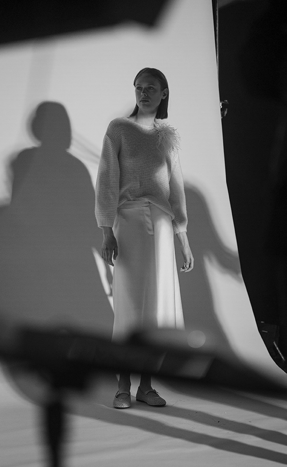 chwarz-weißes Studiofoto der LUISA CERANO Frühling/Sommer 2026: Model mit Bob-Haarschnitt in fedrigem Pullover und langem weißem Rock, mit Schatten und Ausrüstung im Bild