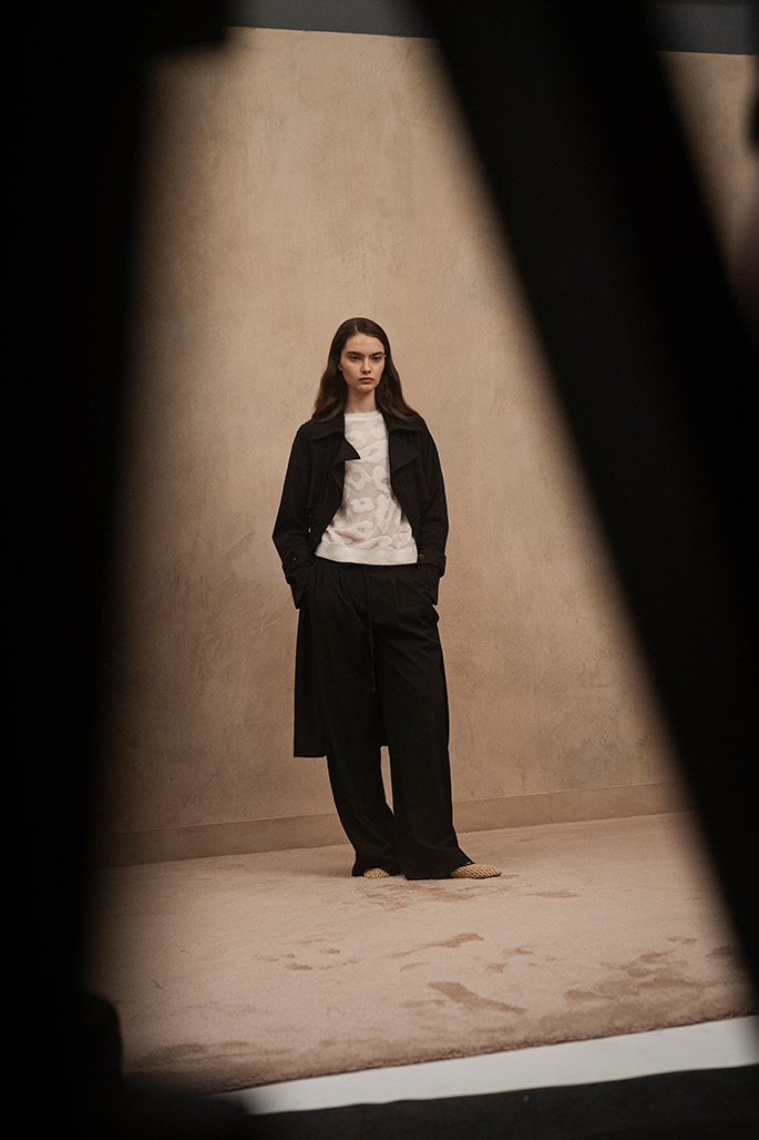 Model in schwarzem Mantel, gemustertem weißem Top und weiten Hosen steht mit Händen in Taschen vor beiger Wand im Fotoshooting