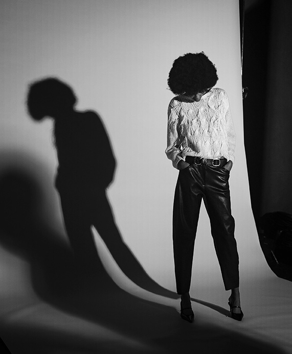 Schwarz-weißes Modefotoshooting der LUISA CERANO Frühling/Sommer 2026 Kampagne: Model mit Afro-Haaren in Spitzenpullover, Lederhose, Gürtel und High Heels wirft dramatischen Schatten im Studio