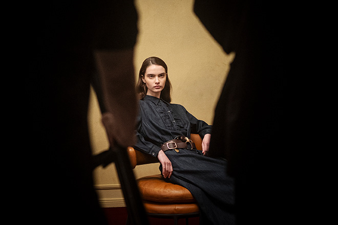 Model mit langen Haaren in dunklem Rüschenkleid und Gürtel sitzt auf Lederstuhl, umrahmt von Schatten im Fotoshooting