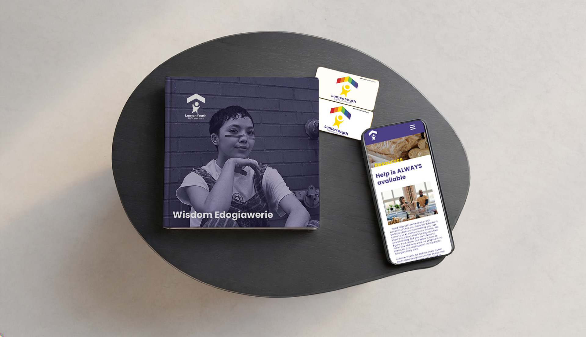 A flat lay mockup displaying the LumenYouth brand identity system, including a hardcover portfolio book, two branded business cards, and a smartphone showing the LumenYouth resources webpage. Brand design by Wisdom Edogiawerie. Mockup template via Adobe Stock and Adobe Firefly.