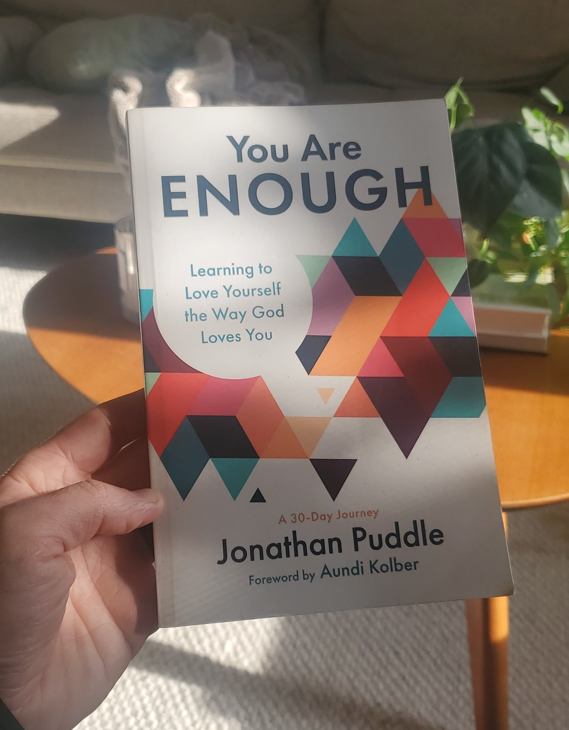 Hand holding the book "You Are Enough: Learning to Love Yourself the Way God Loves You" by Jonathan Puddle, showing the colorful geometric triangle cover design