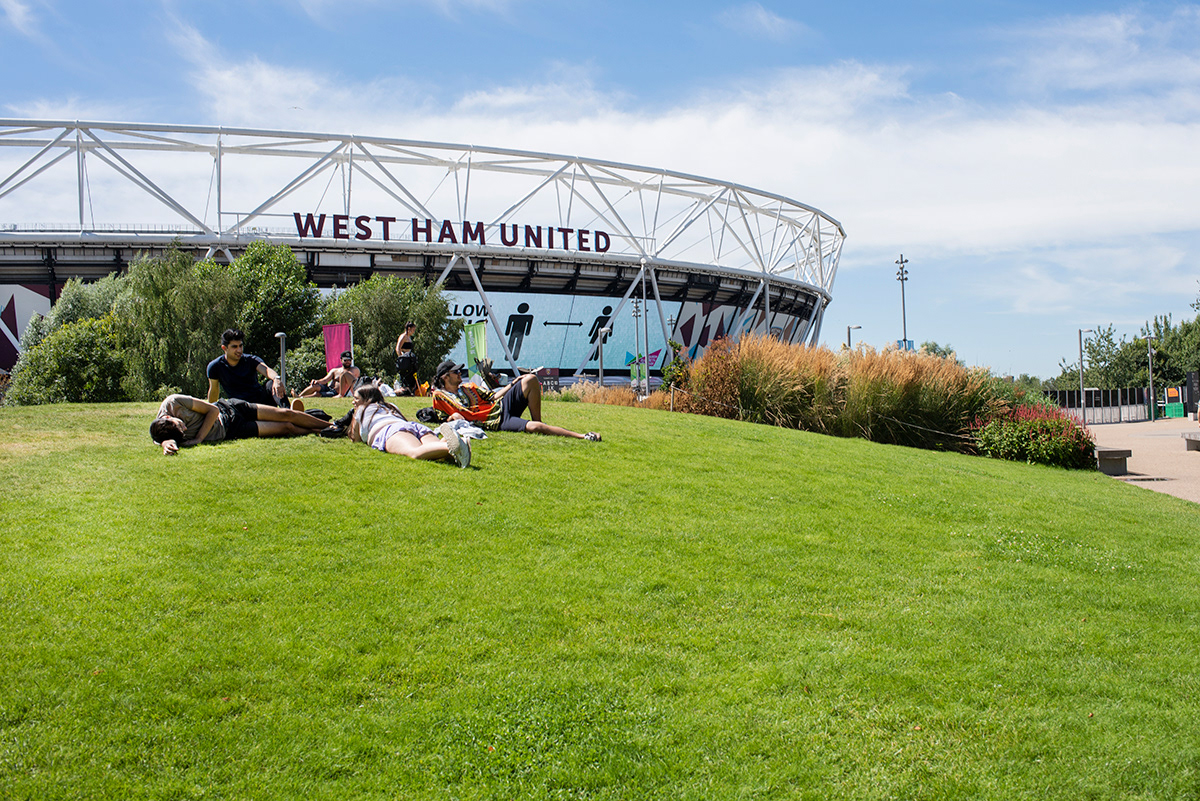 People enjoying the sun in front of the Olympic Stadium, Queen Elizabeth Olympic Park, Stratford, East London.  Signage to encourage social distancing has been placed in every visible outdoor area to prevent mass gathering.