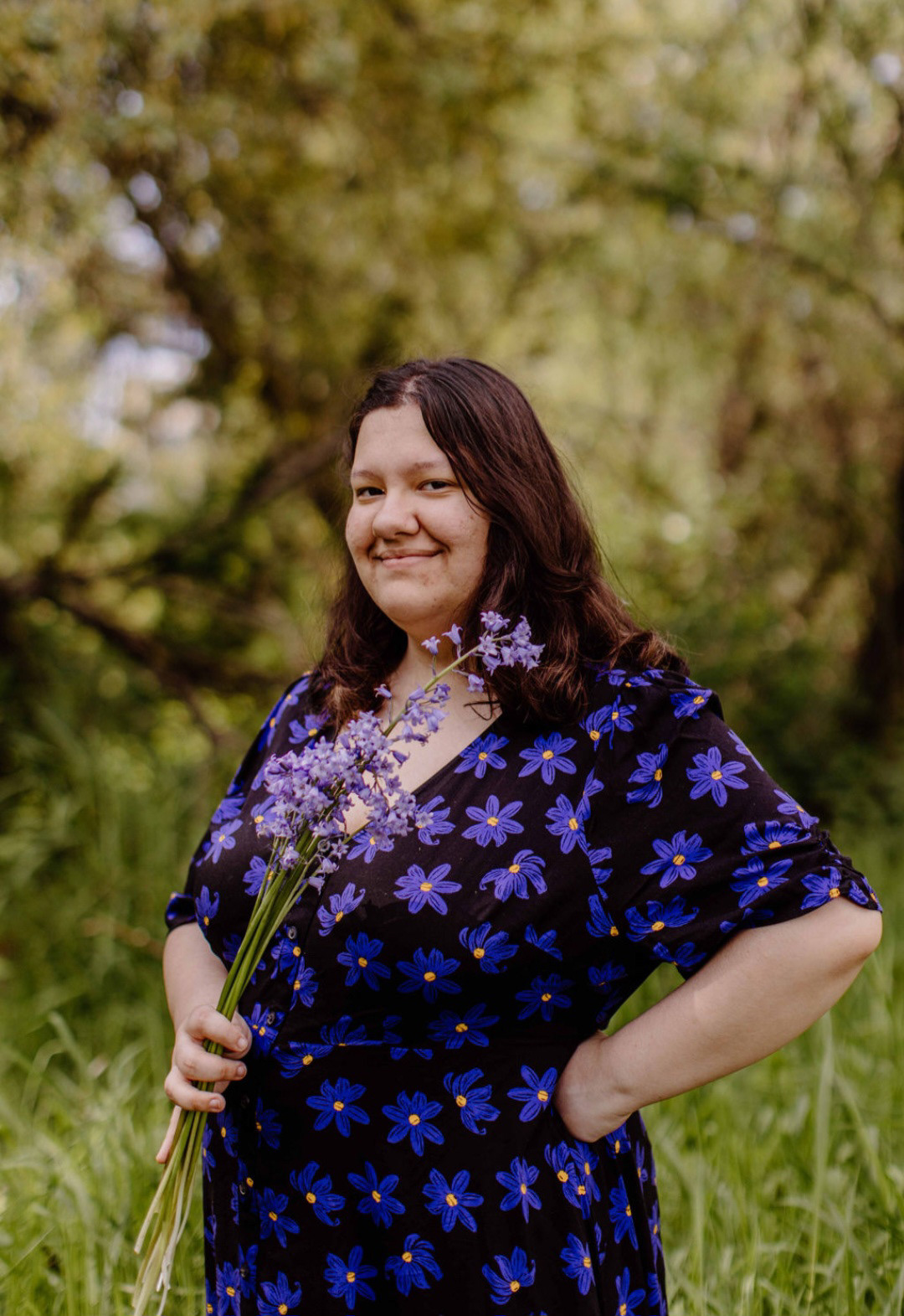 women holding purple flowers in a black dress with dark blue flower pattern with nature in the background.