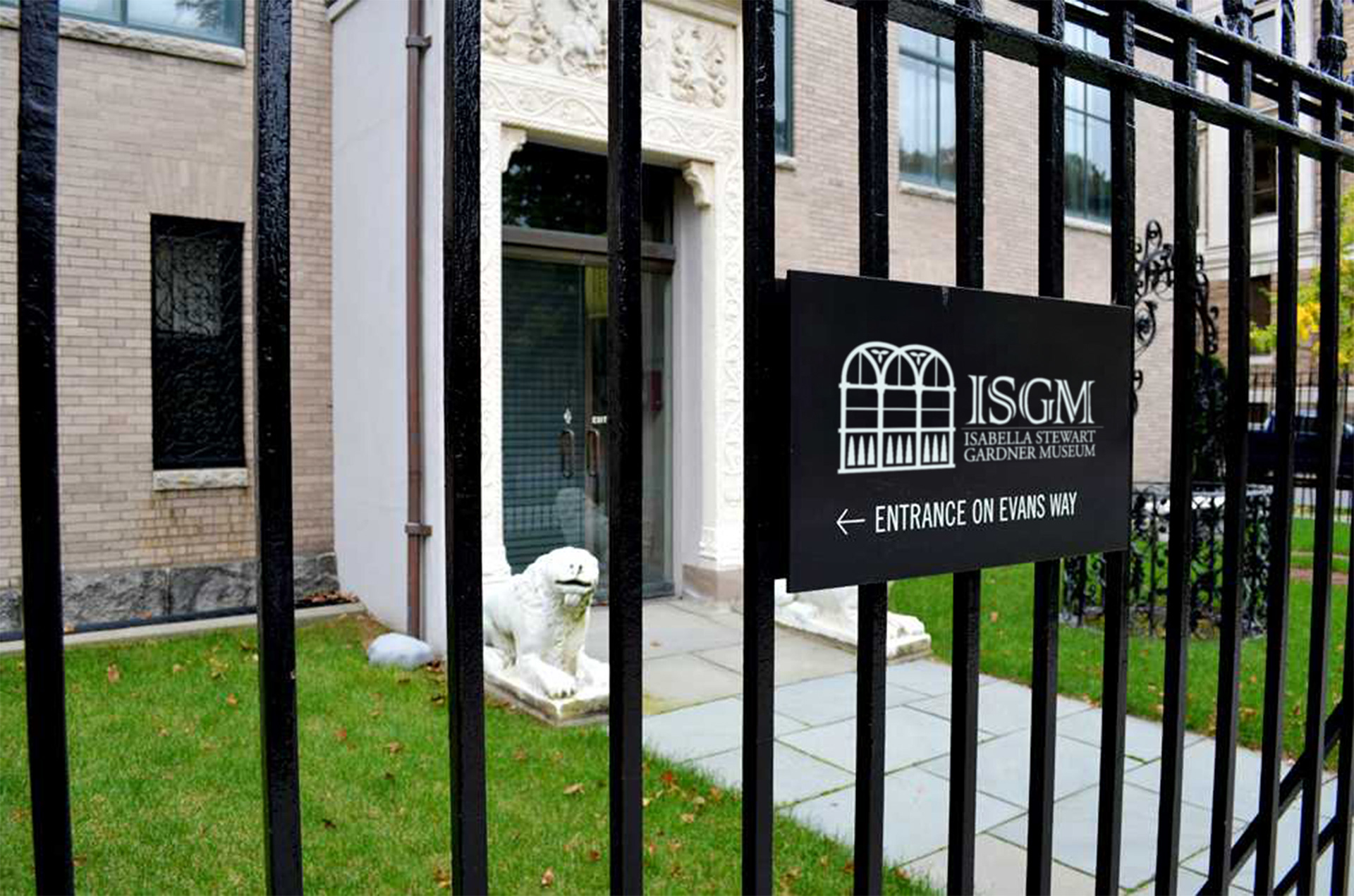 View through a black wrought iron fence with a black sign  and white lettering. The sign shows an updated logo for Isabella Stewart Gardner Museum and an arrow indicating the direction of the entrance.